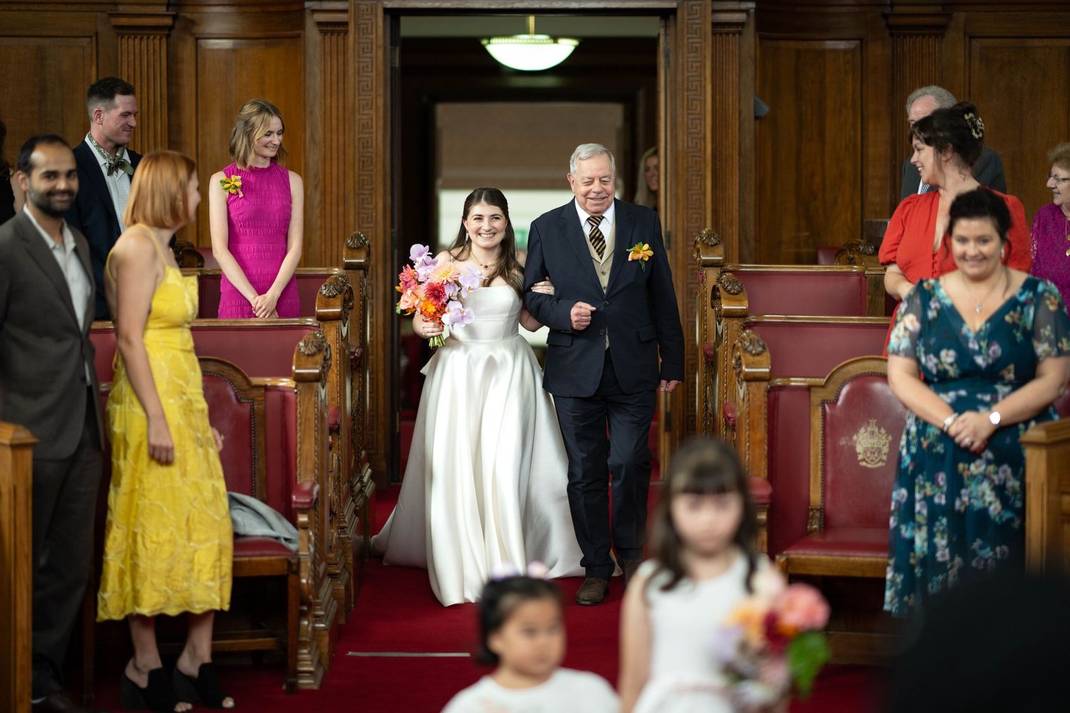 An intimate wedding ceremony takes place inside a historic wood-paneled chapel with attendants standing along the red-carpeted aisle.