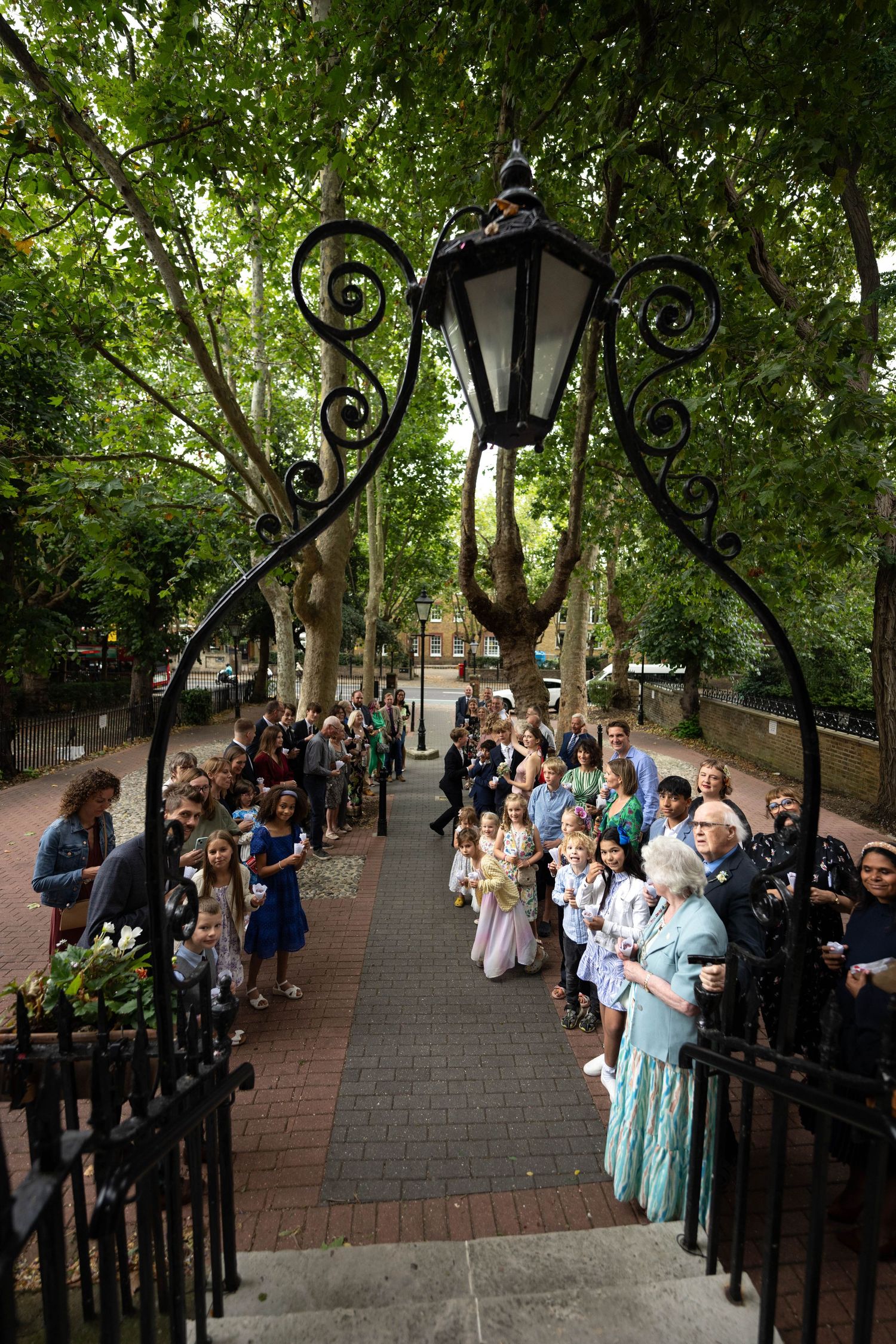 A crowd gathers along a brick pathway with ornate black lamp posts and iron railings under a canopy of green trees.