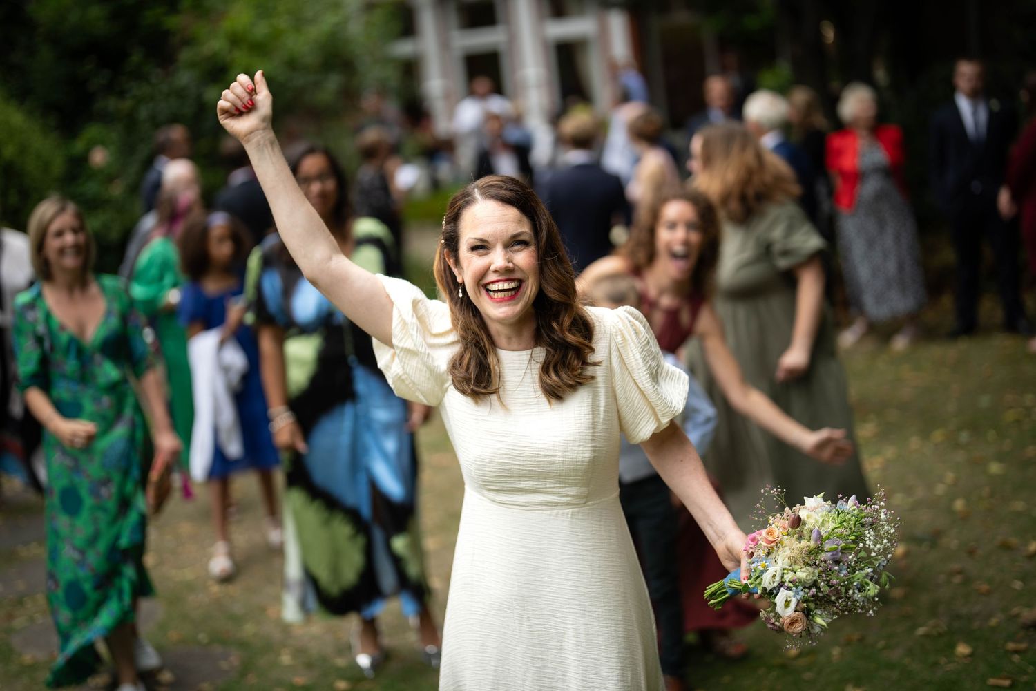 Bride in white dress holds bouquet and raises arm triumphantly with wedding guests celebrating in background.
