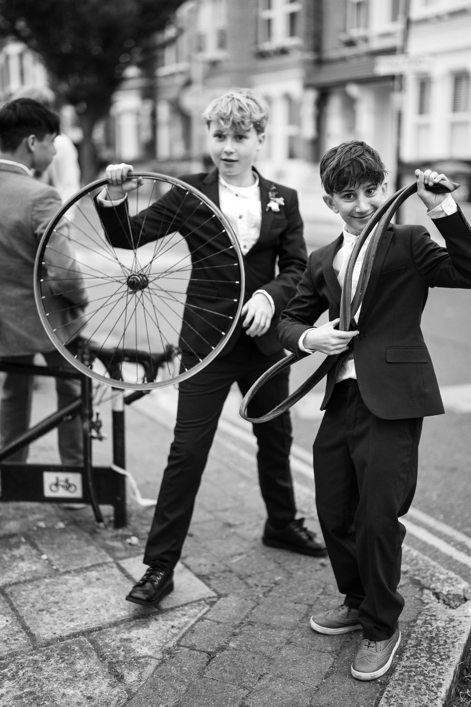 Black and white vintage photo of young boys in suits examining bicycle wheels on a city street.