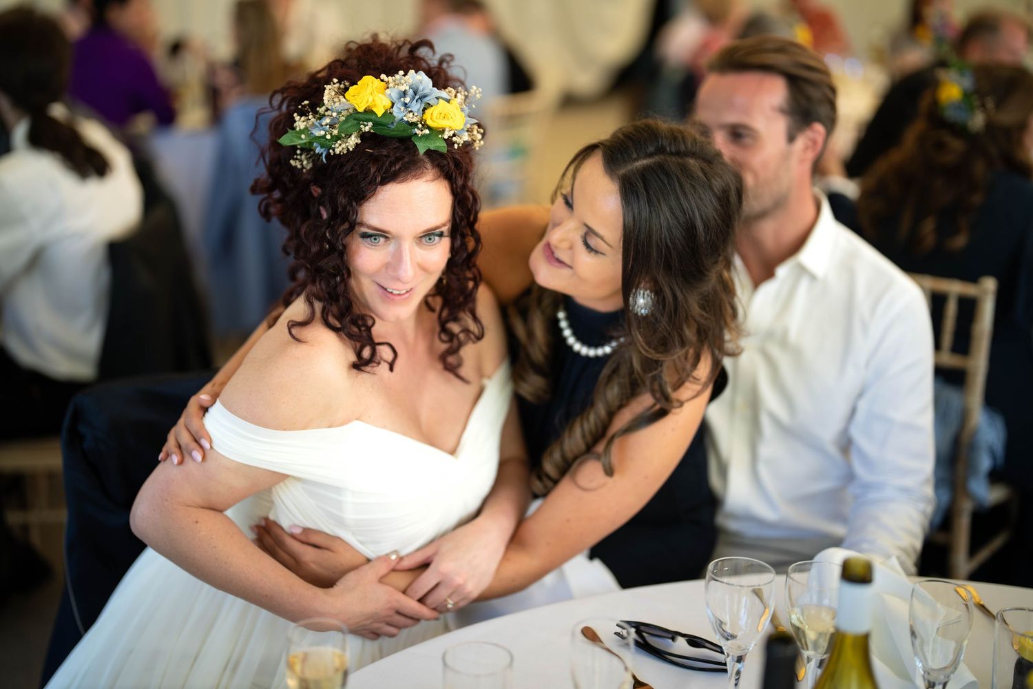 Two friends share a joyful moment at a wedding reception as one wears a white dress with floral crown.