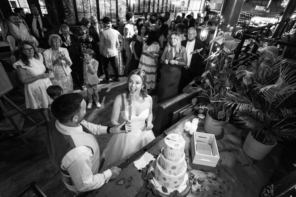 A black and white celebration shows a three-tiered wedding cake being cut while guests gather around in an indoor venue.