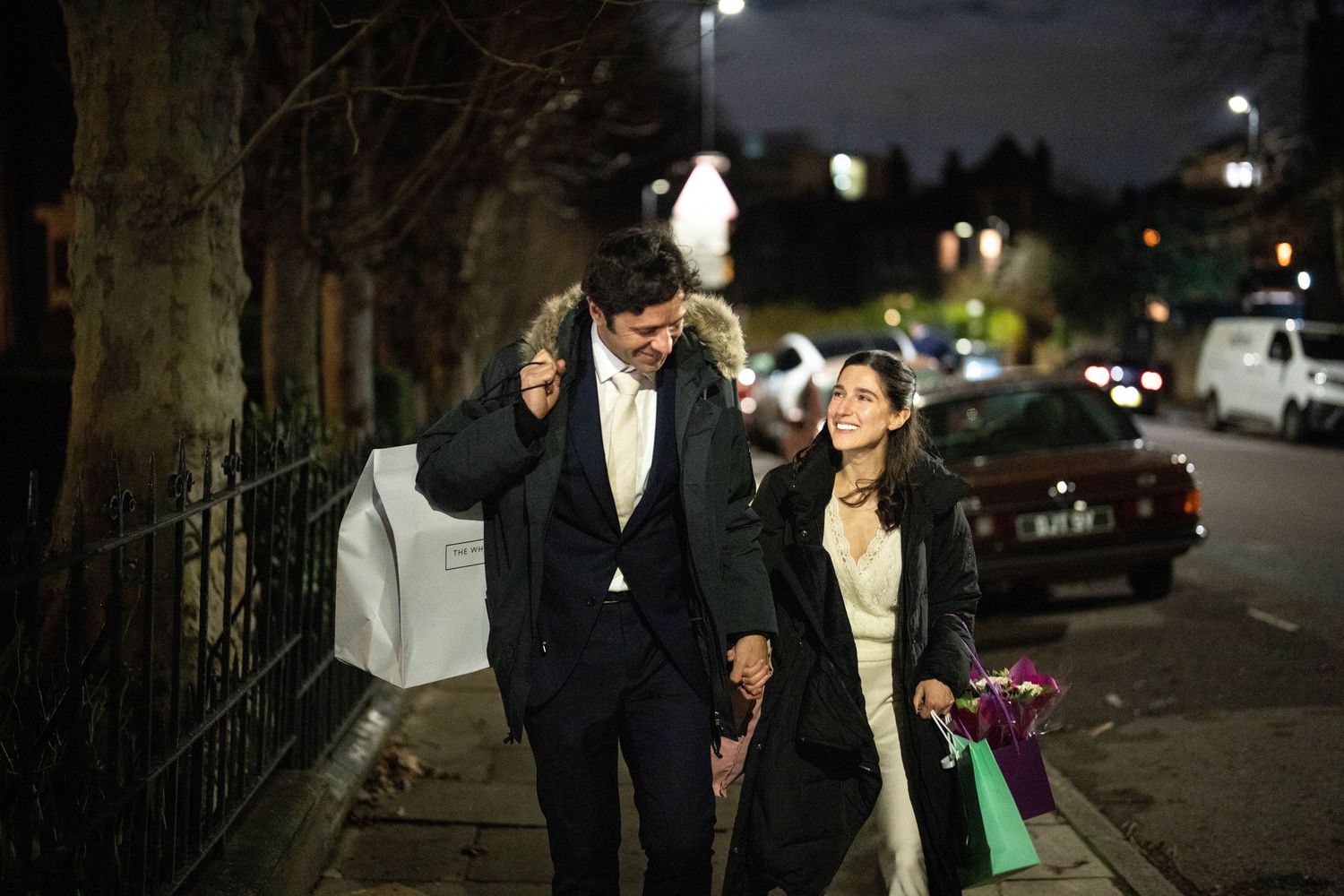 A couple dressed in formal attire walks down a nighttime city street while carrying shopping bags.