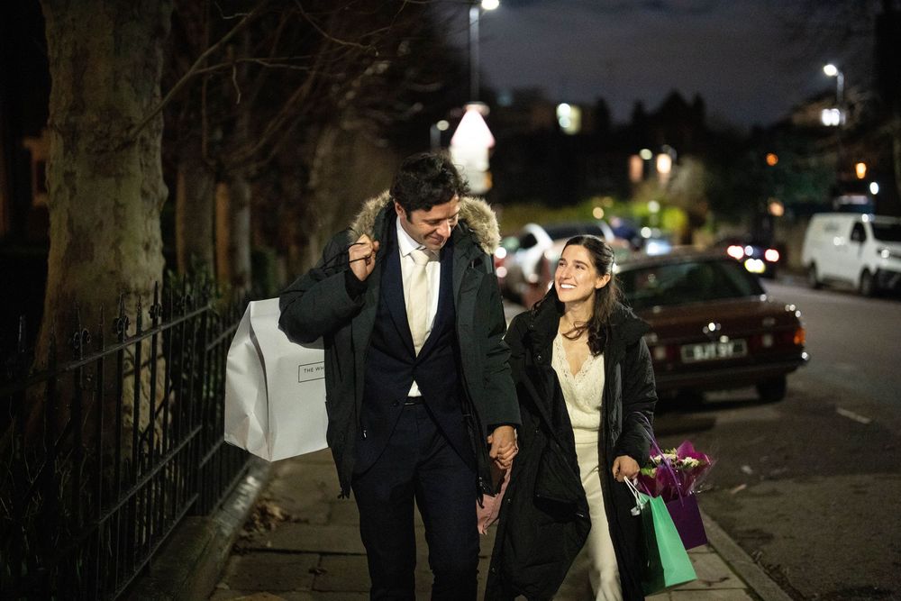 A couple dressed in formal attire walks down a nighttime city street while carrying shopping bags.