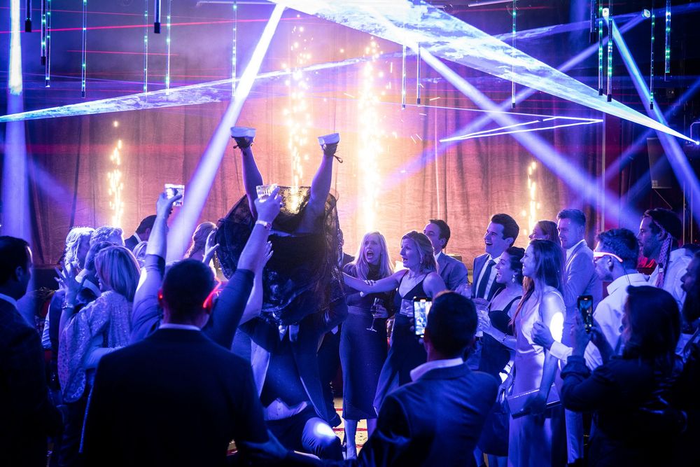 Party crowd dances and raises hands under blue and purple laser lights at a nightclub event.