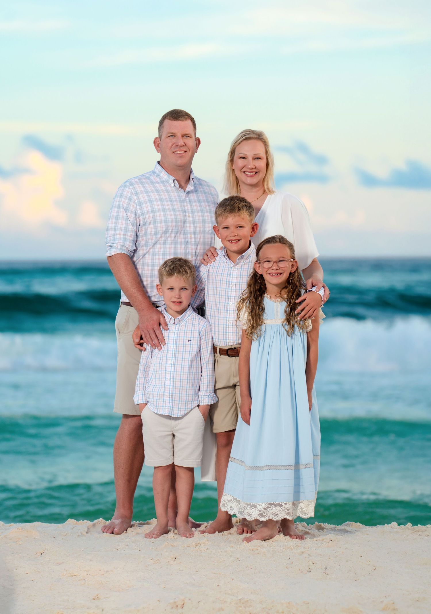 Family wearing coordinated neutral outfits poses together on wooden boardwalk with palm trees in background.