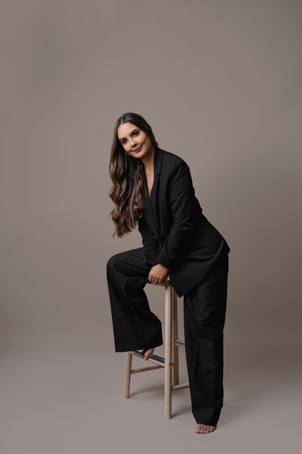 Fashion portrait of woman in black pantsuit seated on wooden stool against neutral background.