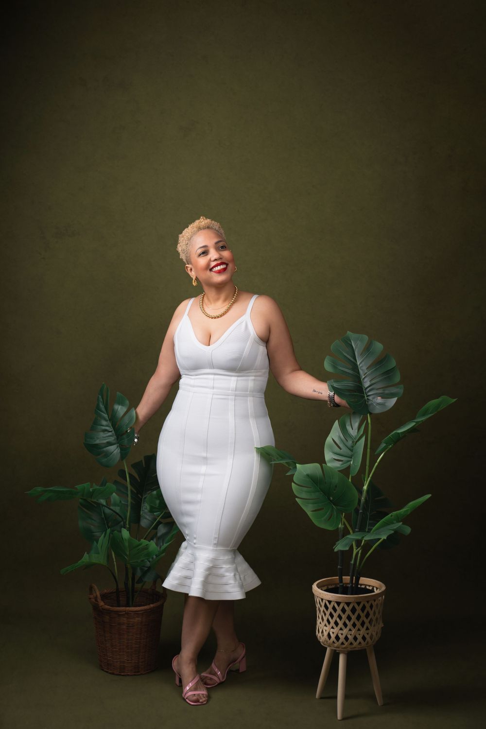 Person in white dress poses between potted monstera plants against dark green backdrop.