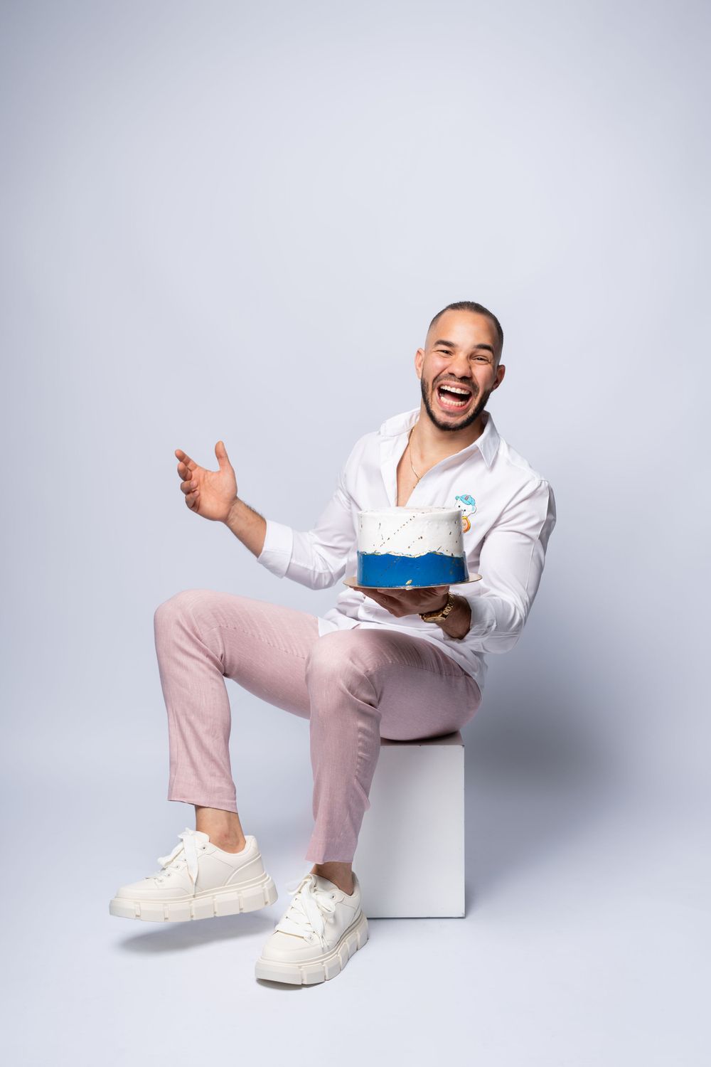 Energetic studio portrait of person celebrating with cake while seated on white cube.