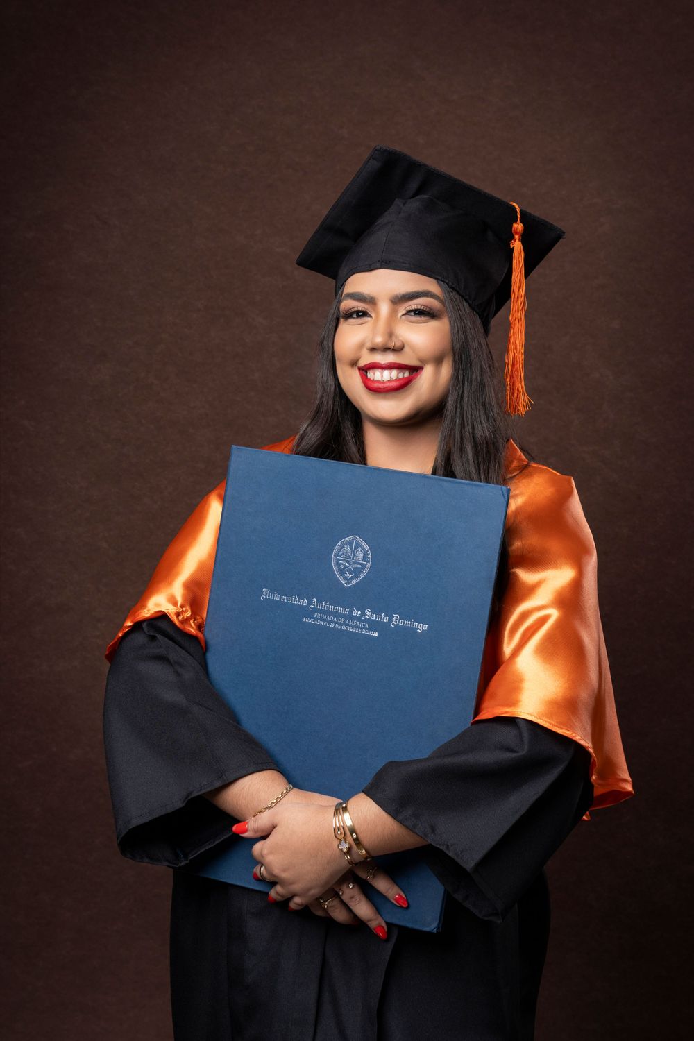 Graduate in orange and black regalia proudly displays diploma against brown background.