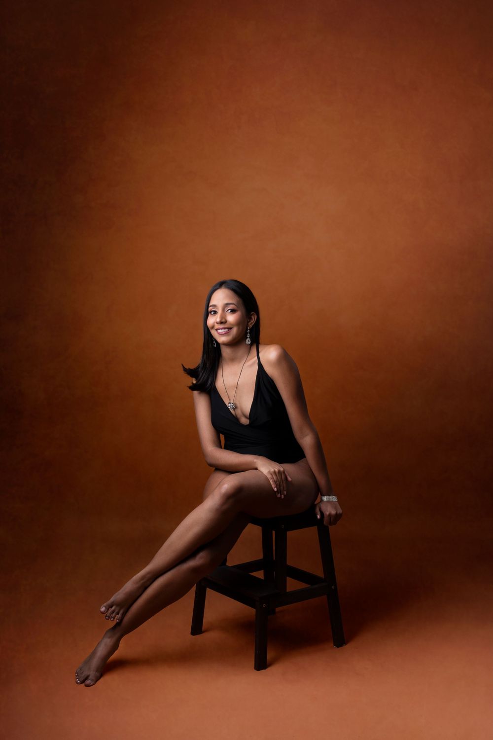 Elegant portrait of person in black dress posed on stool against warm brown background.