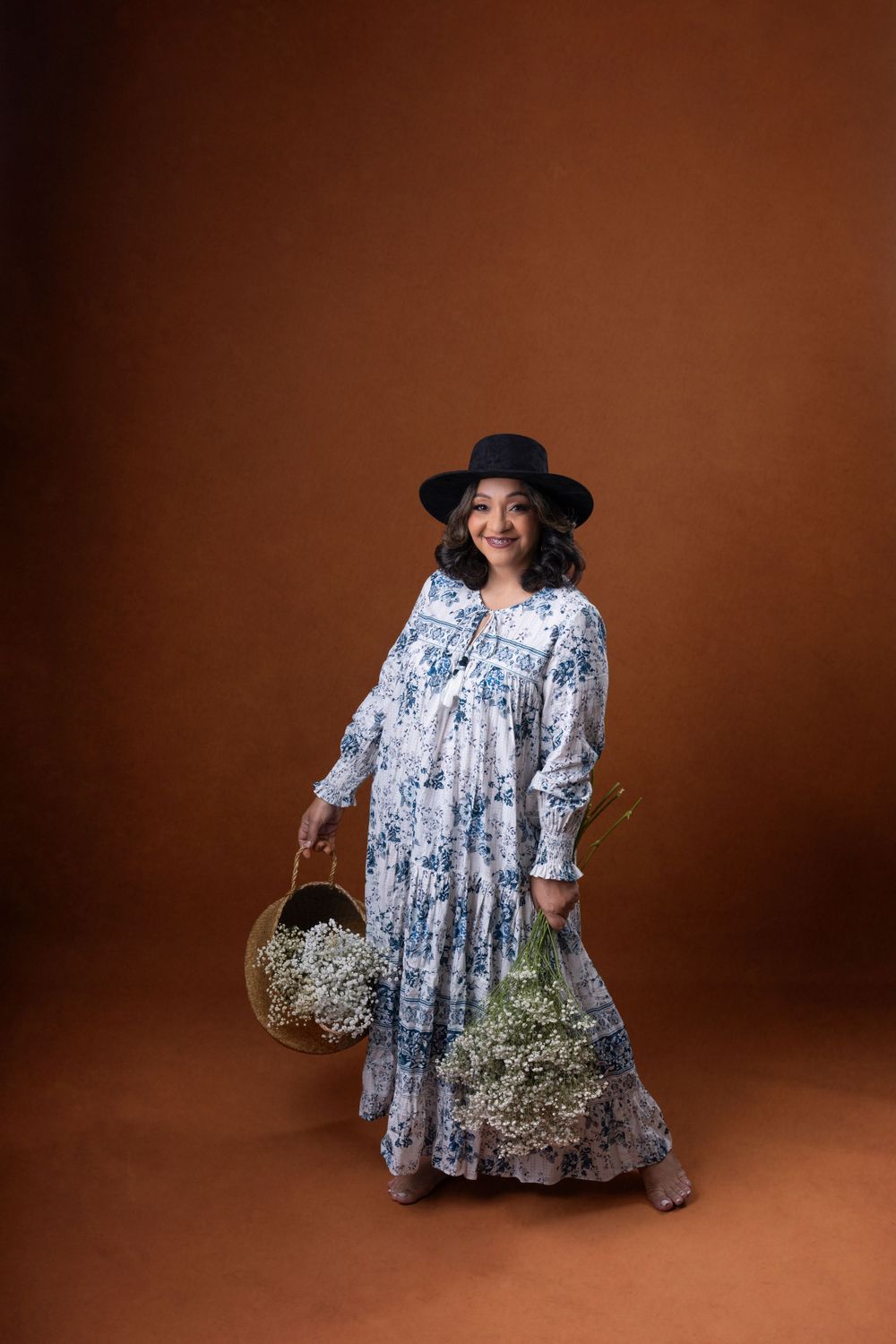 Person in flowing blue dress and hat holds basket against brown studio background.