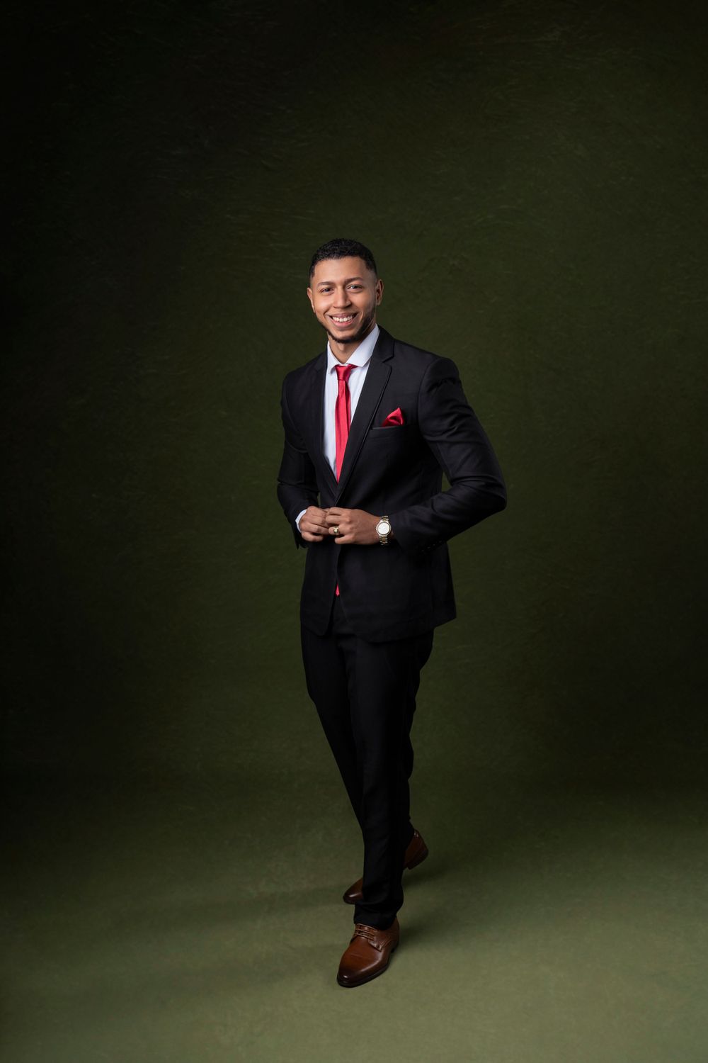 Professional in black suit with red tie poses against dark green background.