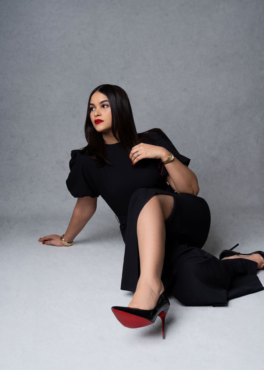 Fashion portrait of woman in black dress with red-soled shoes against grey background.