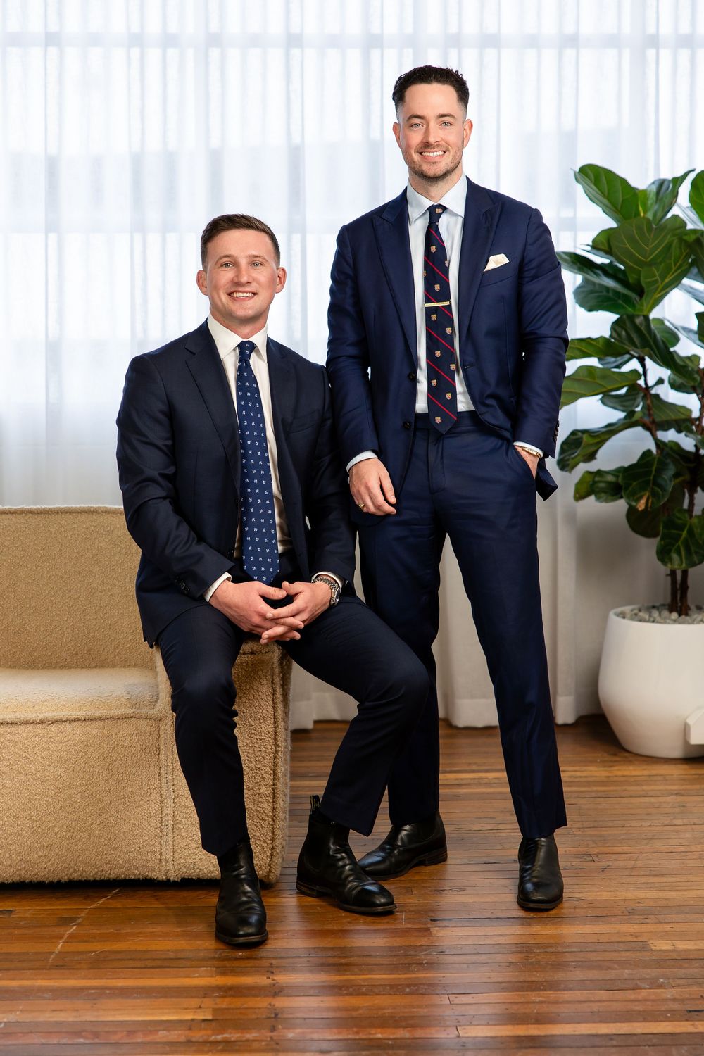 Two businessmen in navy suits and blue ties sit casually on hardwood floor with potted plant in background.