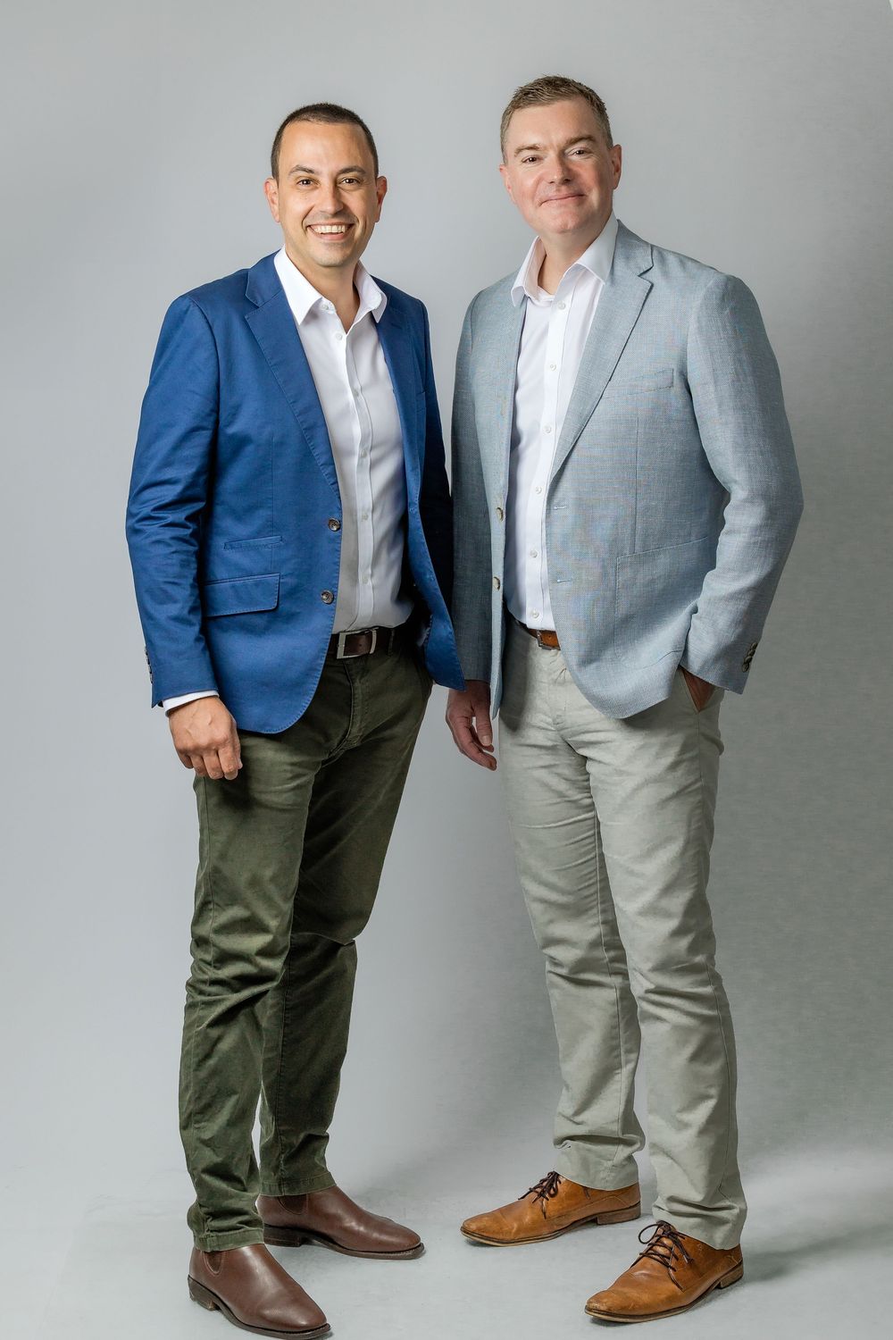 Two people in business attire stand together against a gray backdrop wearing blue blazer and gray suit.