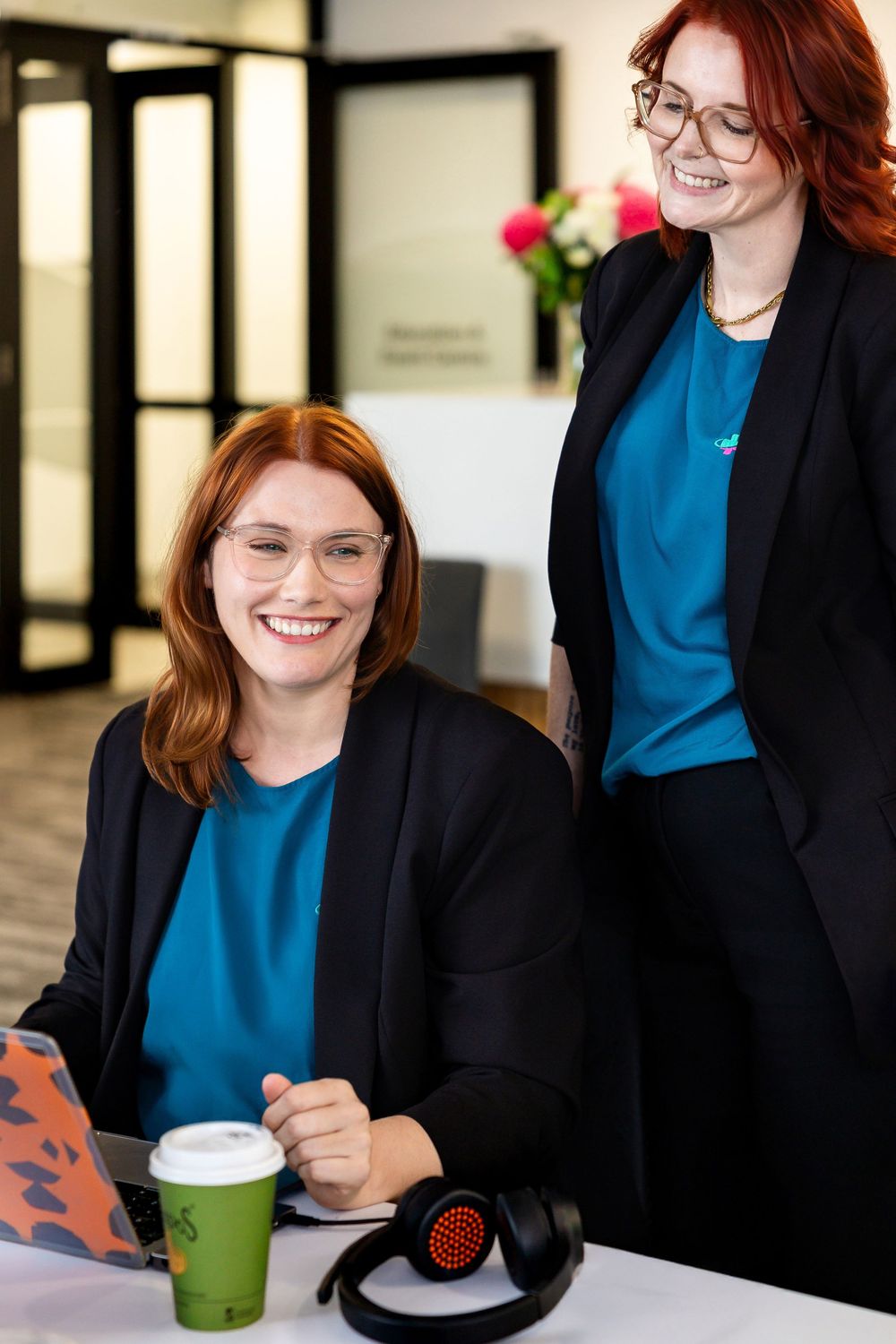 Professional in a black blazer and blue top smiles while working at a desk with a green coffee cup.