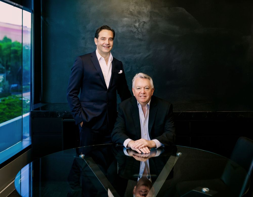 Two businessmen in formal attire pose in a modern office setting with dark walls and window lighting.