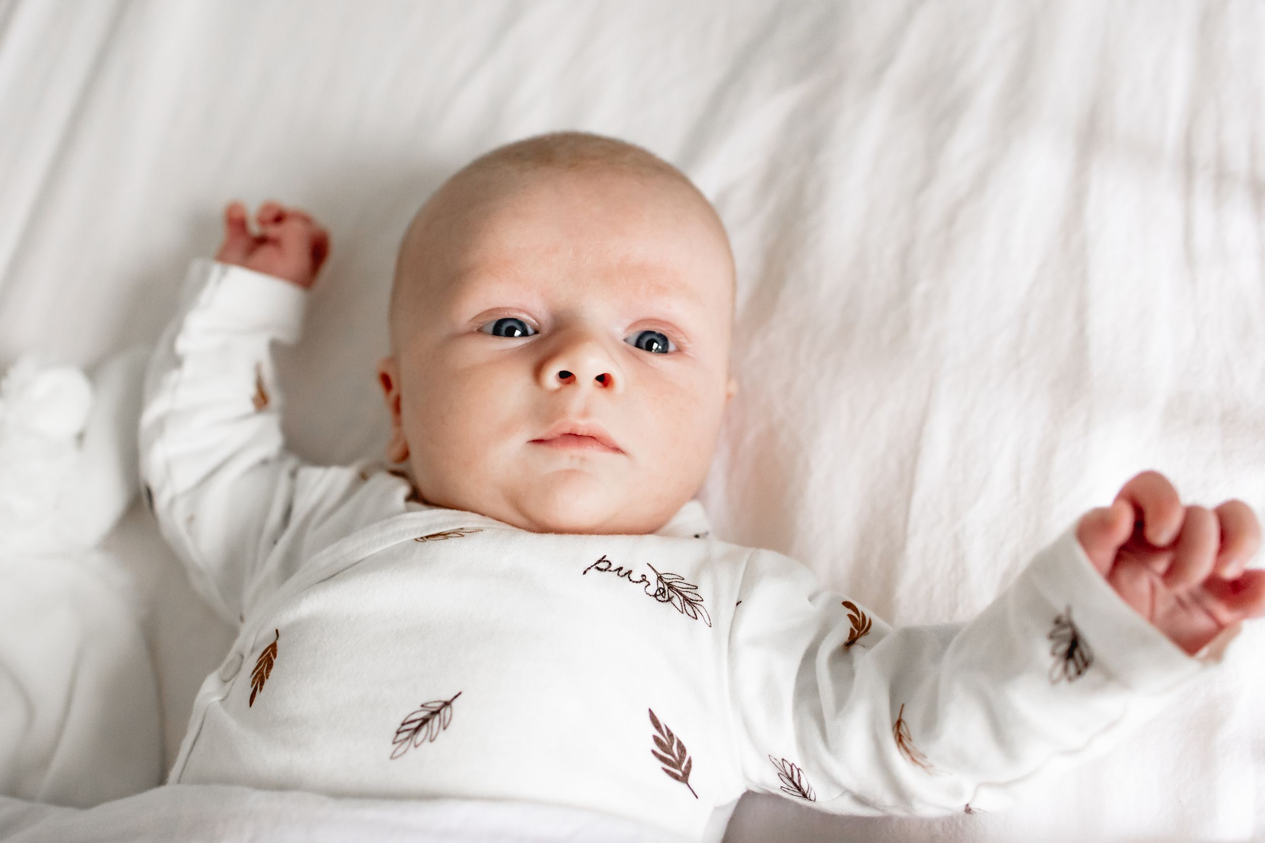 Newborn baby wearing white outfit with feather pattern lying on white bedding.
