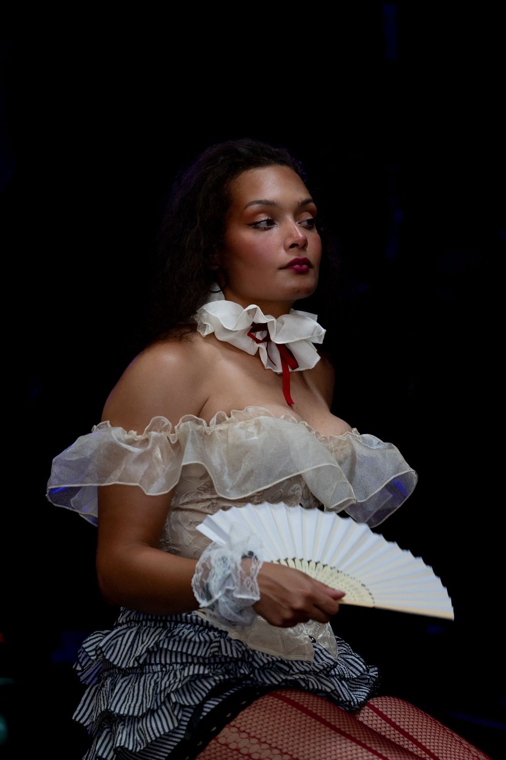 A young woman in a vintage off-shoulder white ruffled costume holds a hand fan against a dark background.