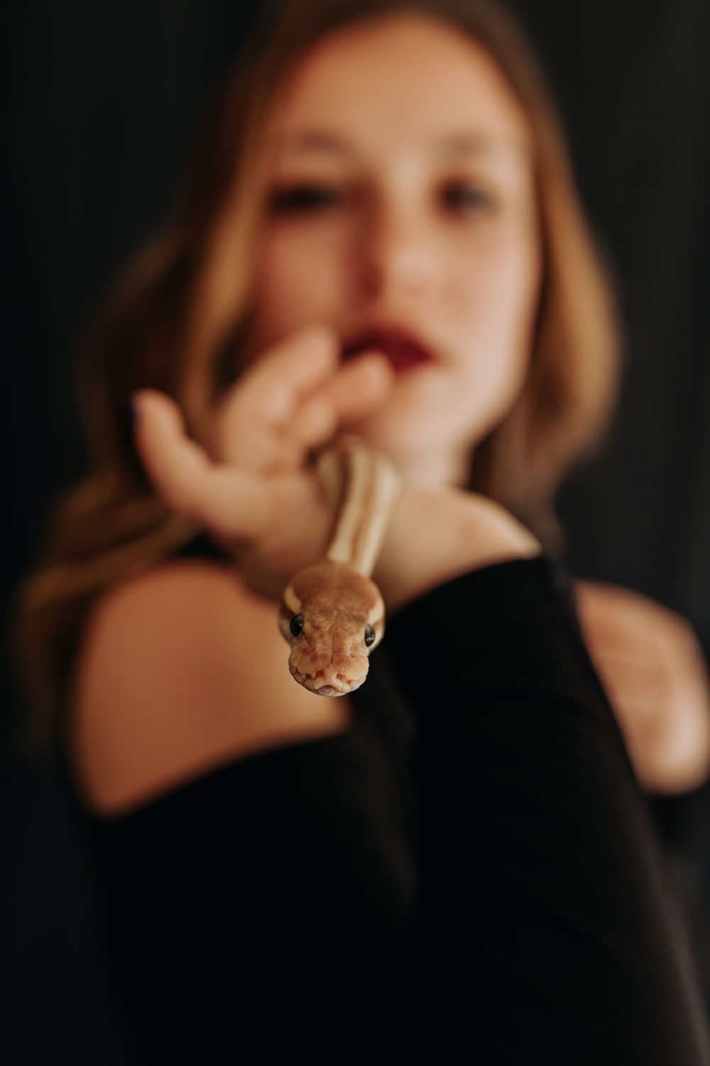 Young woman in black dress holds a small tan snake close to her face against dark background.
