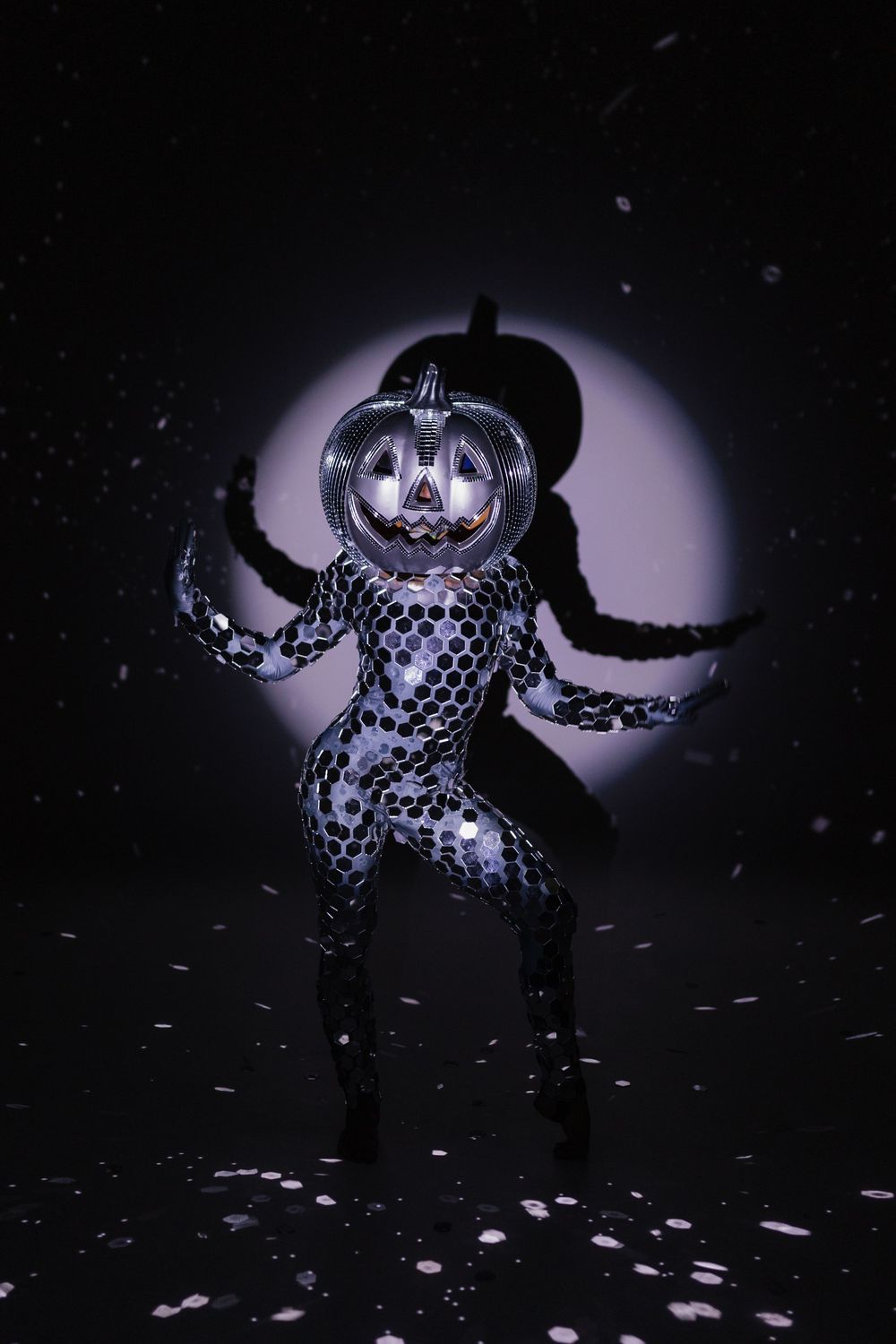 A performer in a metallic pumpkin head costume and sequined bodysuit poses dramatically under a spotlit backdrop.