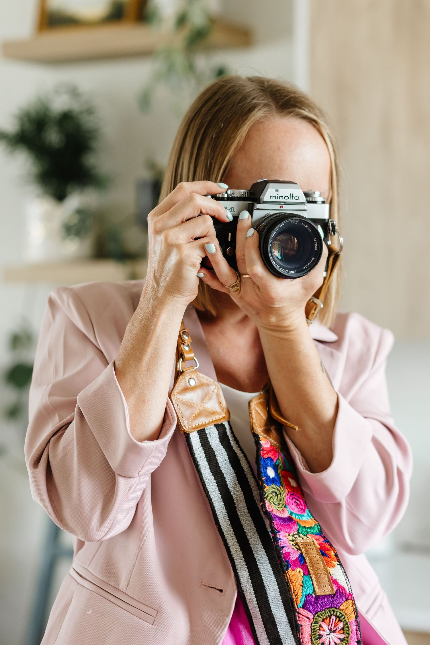 Woman in pink blazer photographing with a vintage Minolta film camera and colorful embroidered strap indoors.