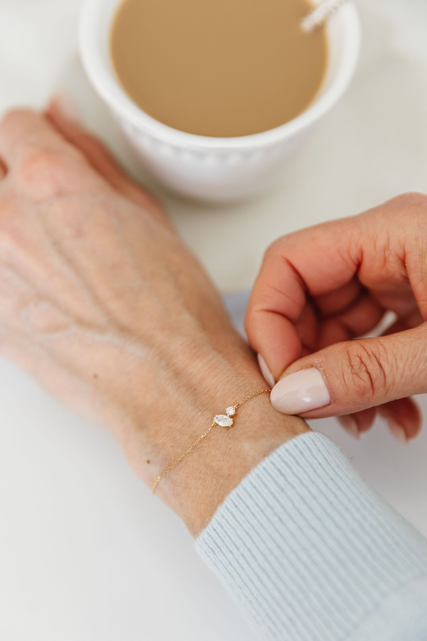 Close-up of delicate gold chain bracelet with heart charm being clasped on wrist near coffee cup.