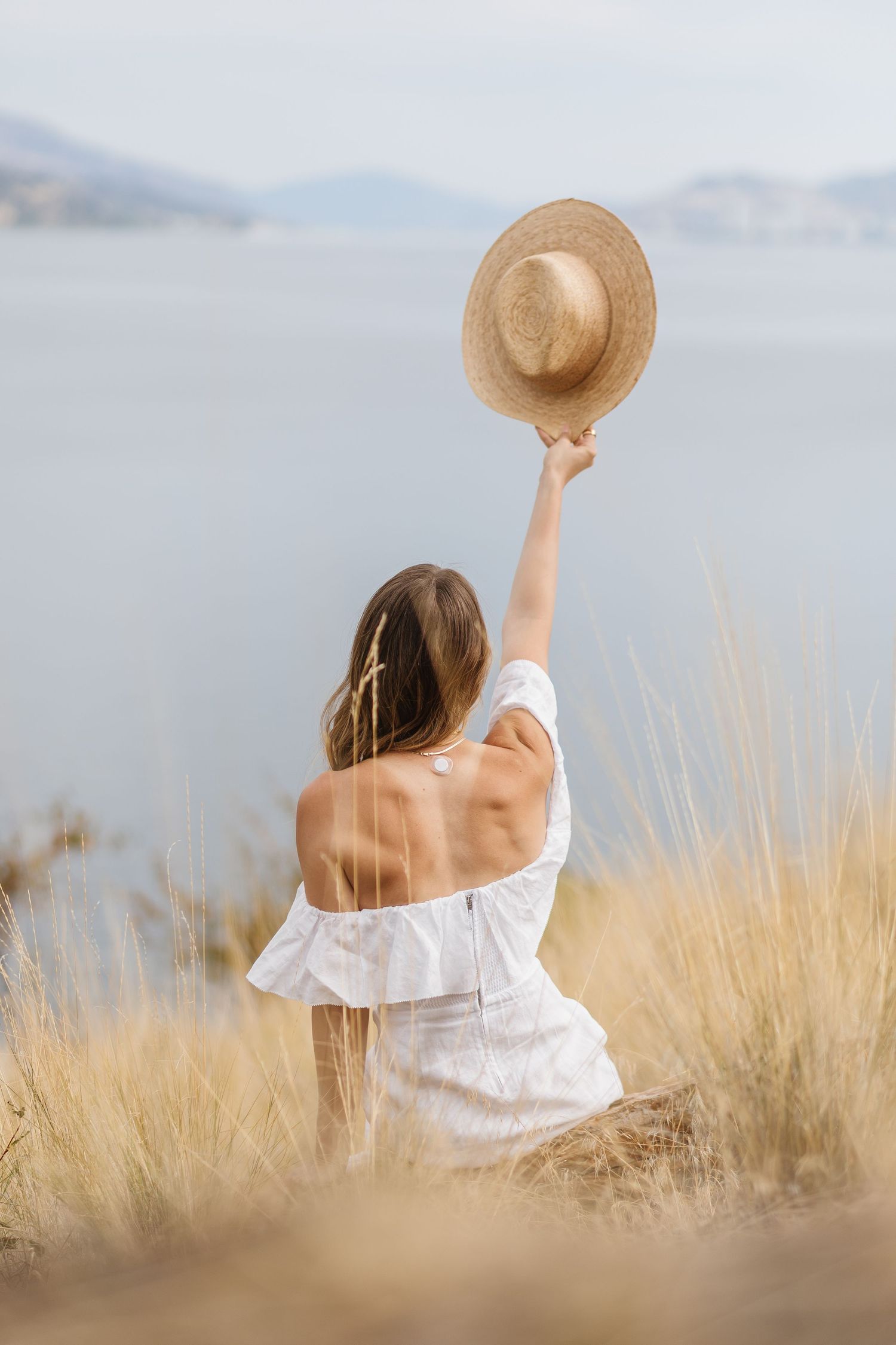 A figure in a white dress and straw hat sits peacefully among tall golden grass overlooking a serene mountain landscape.