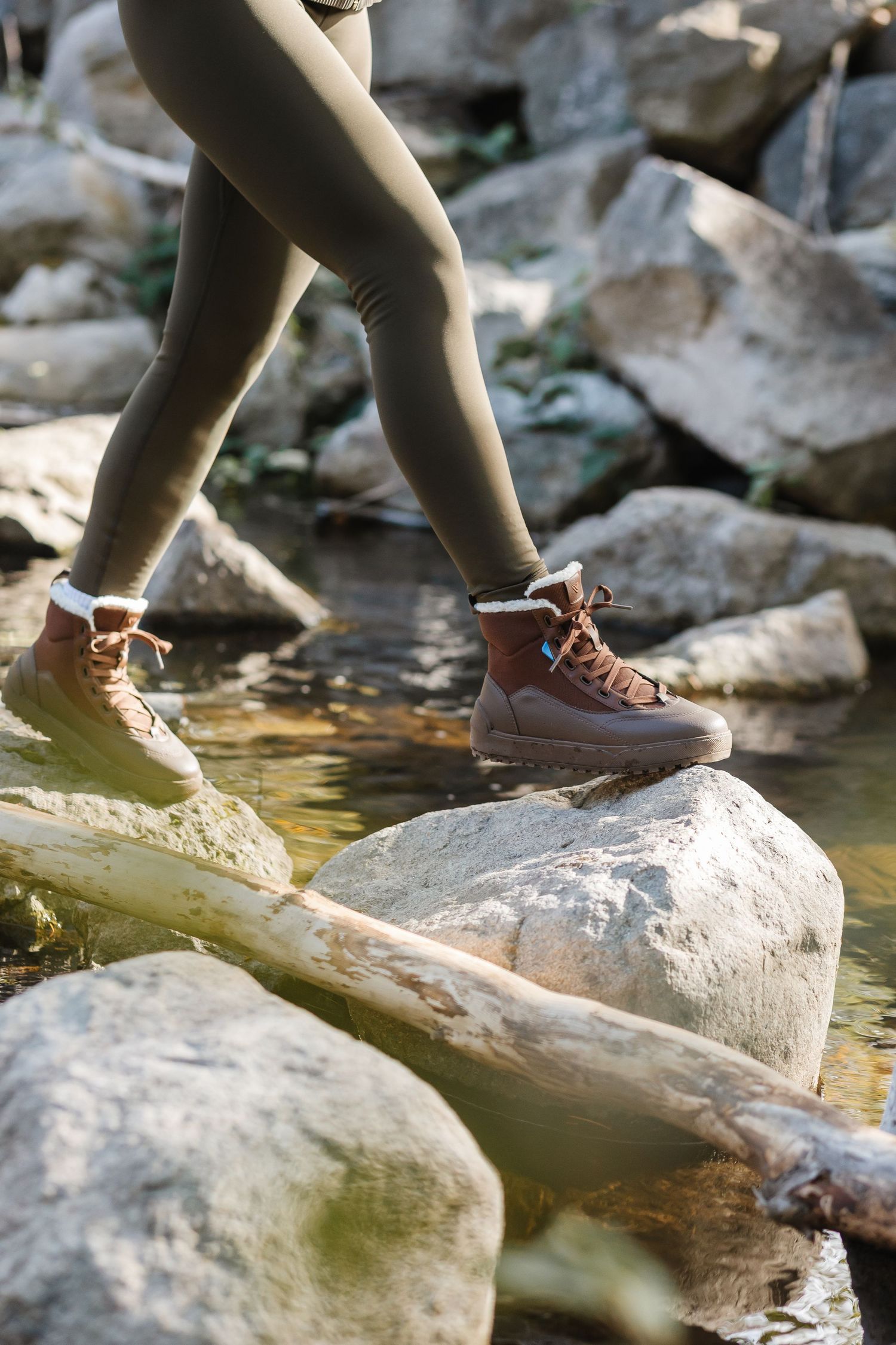 Person stepping on rocks over water wearing brown lace-up hiking boots and olive green leggings in nature.