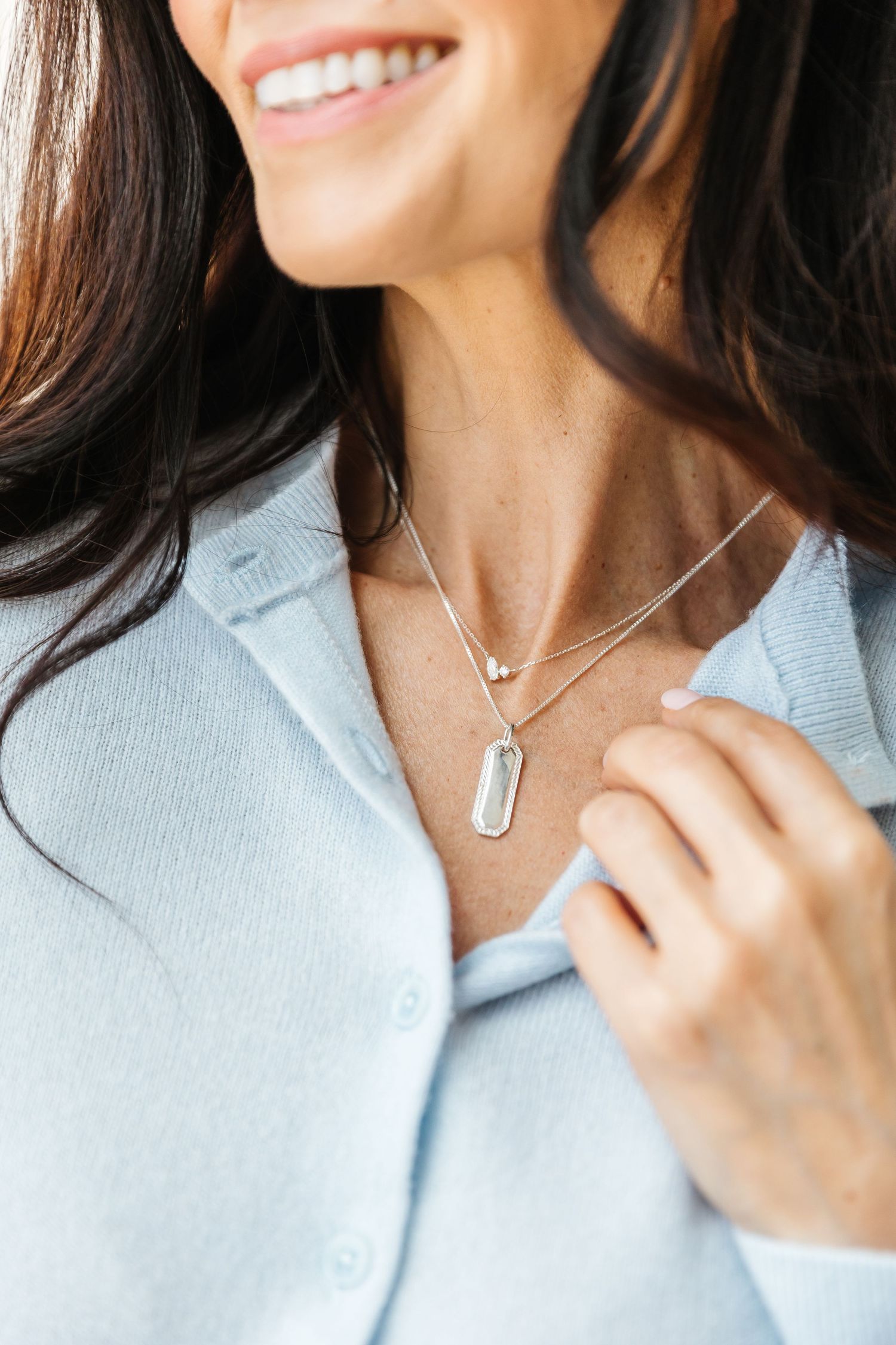 Smiling woman wearing layered silver necklaces with pendant charms, dressed in a light blue button-up cardigan.