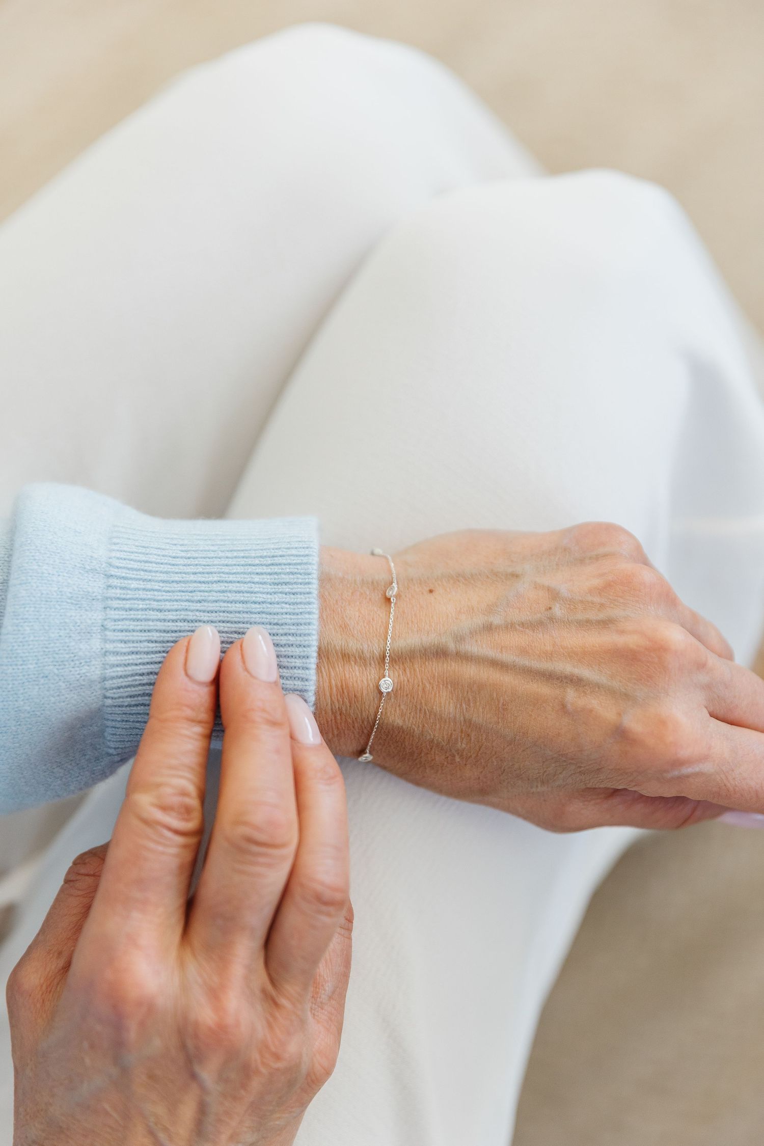 Elderly woman wearing a delicate silver bracelet on her wrist, dressed in a light blue sweater and white pants.