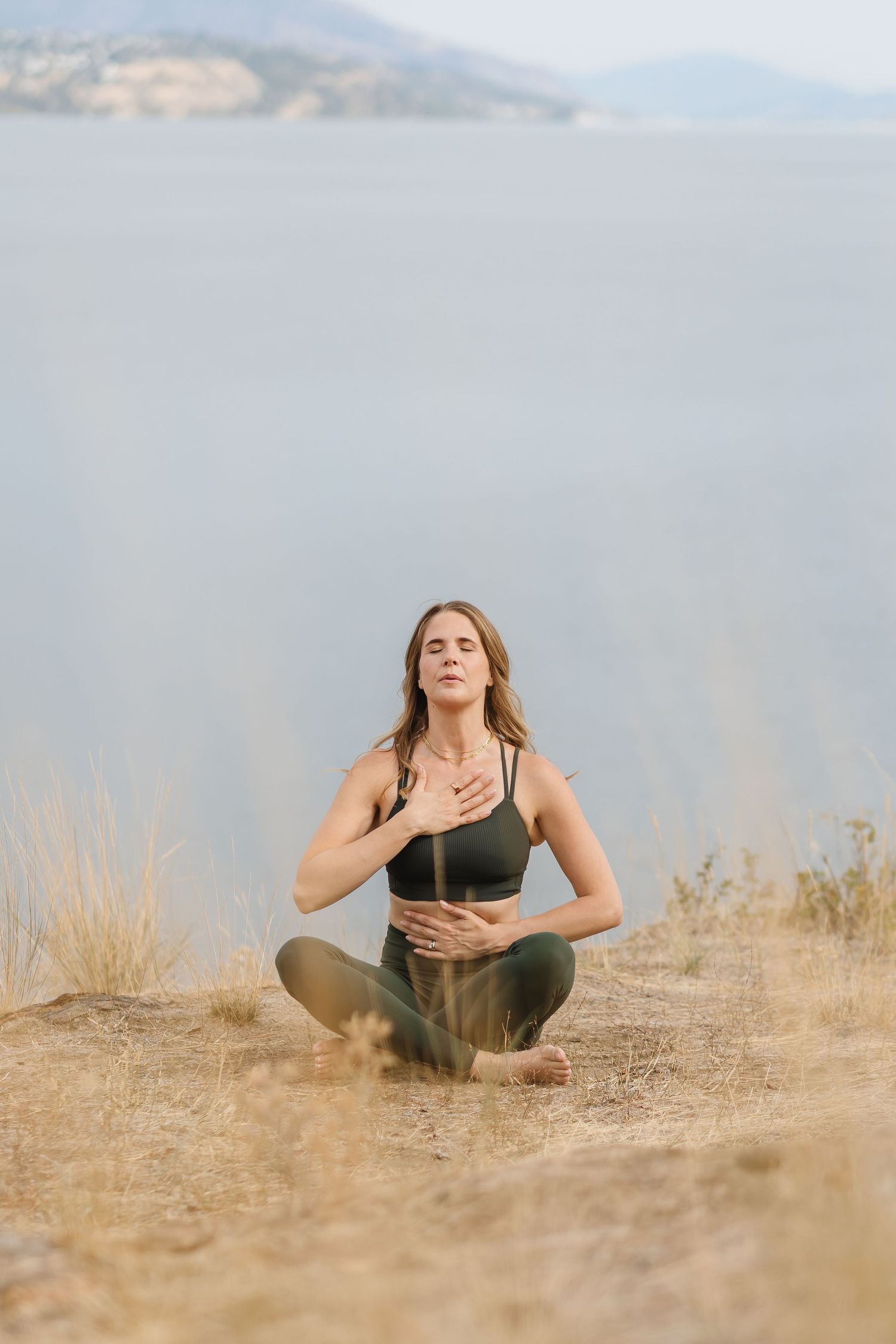 Person in athletic wear doing stretching exercises on dry grass with mountains in the background.