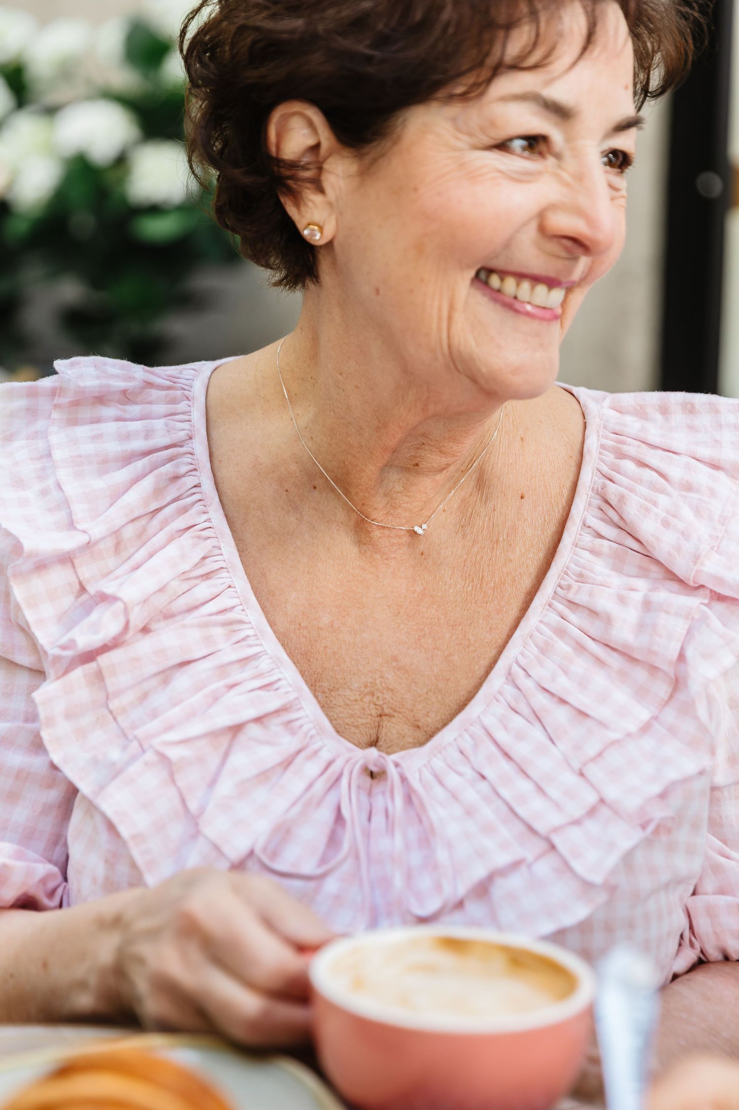 Smiling older woman in a pink ruffled blouse wearing a delicate necklace, enjoying a latte at a cafe.