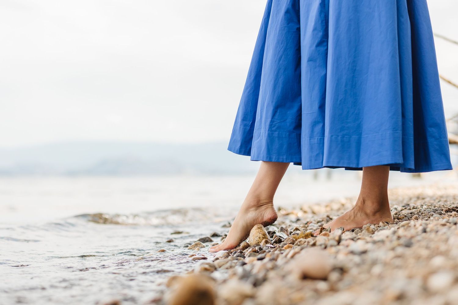 Woman in a flowing blue dress walks barefoot along a rocky beach shoreline at sunset.