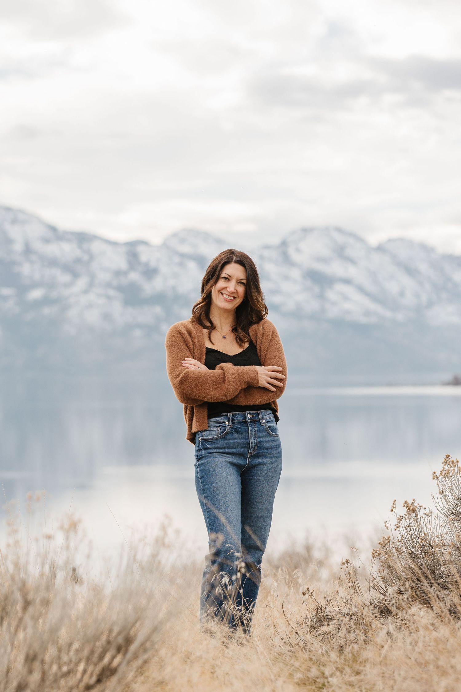 A smiling brunette woman in jeans and brown cardigan poses arms crossed near a lake with snow-capped mountains behind her.