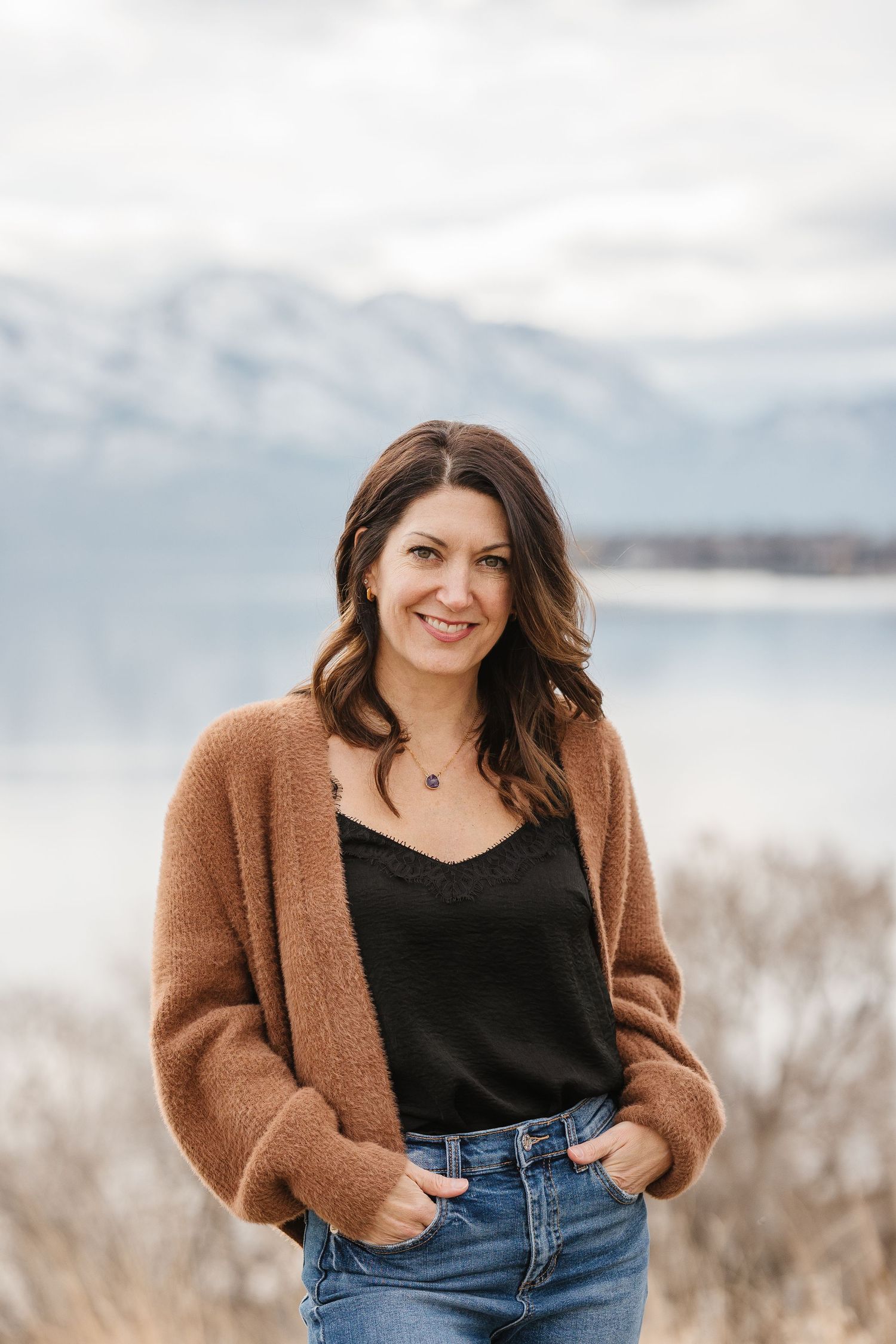 Woman in brown cardigan and jeans smiling outdoors with snowy mountains and lake in background.