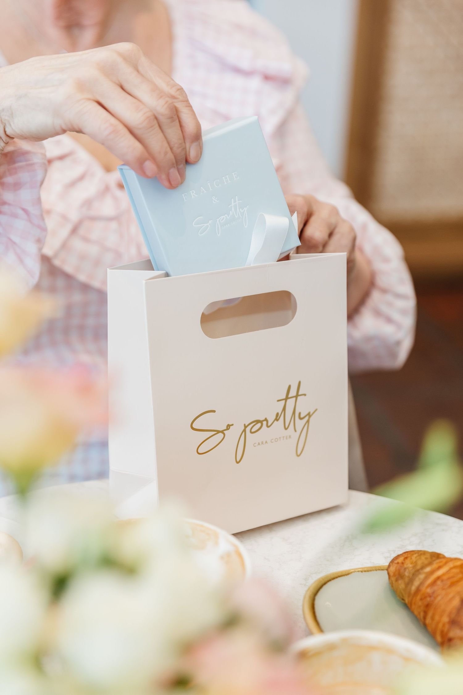 Hands placing blue card into white So Pretty gift bag near croissant and flowers.