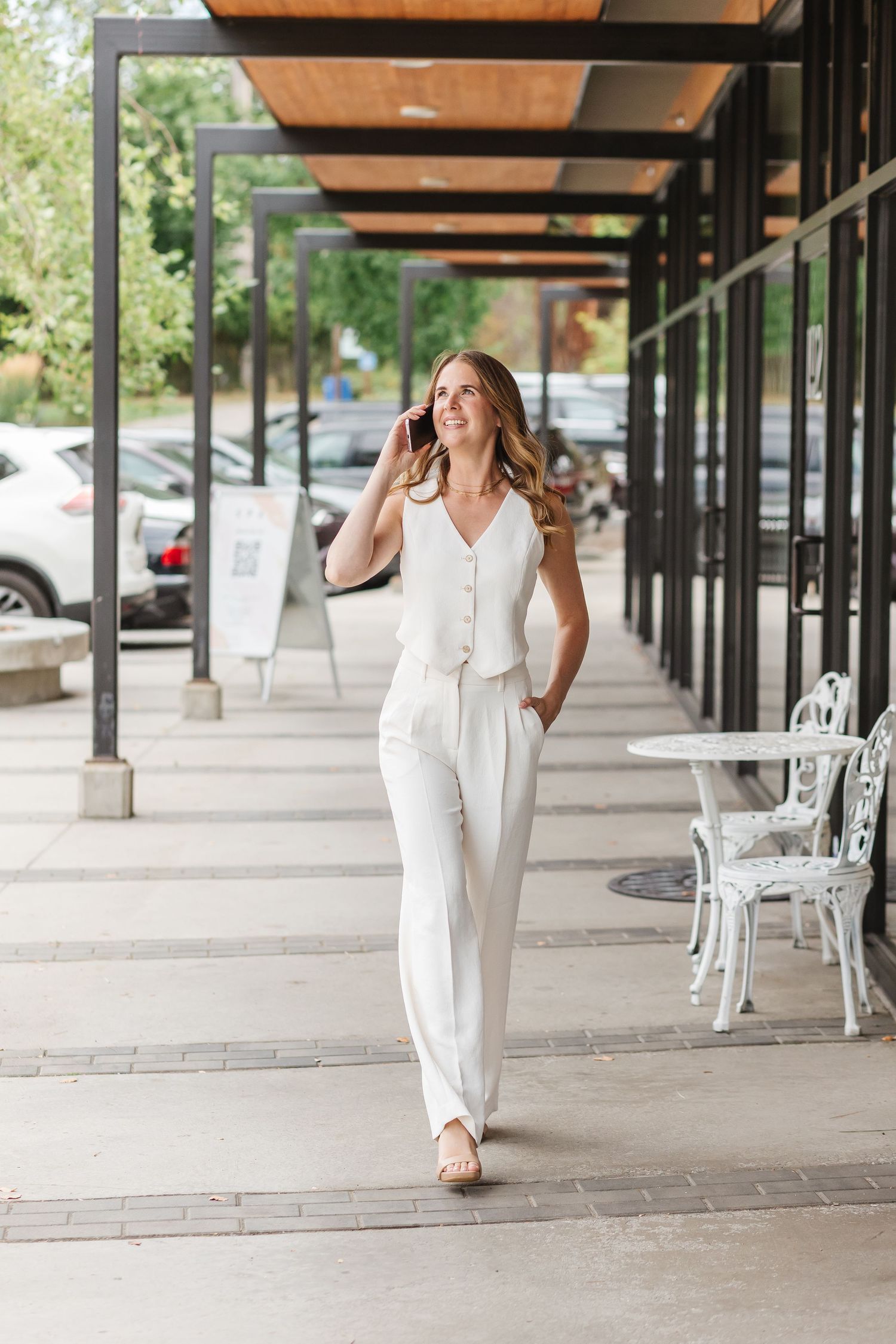 A person in a white pantsuit walks under a covered outdoor walkway with wooden beams and white columns.