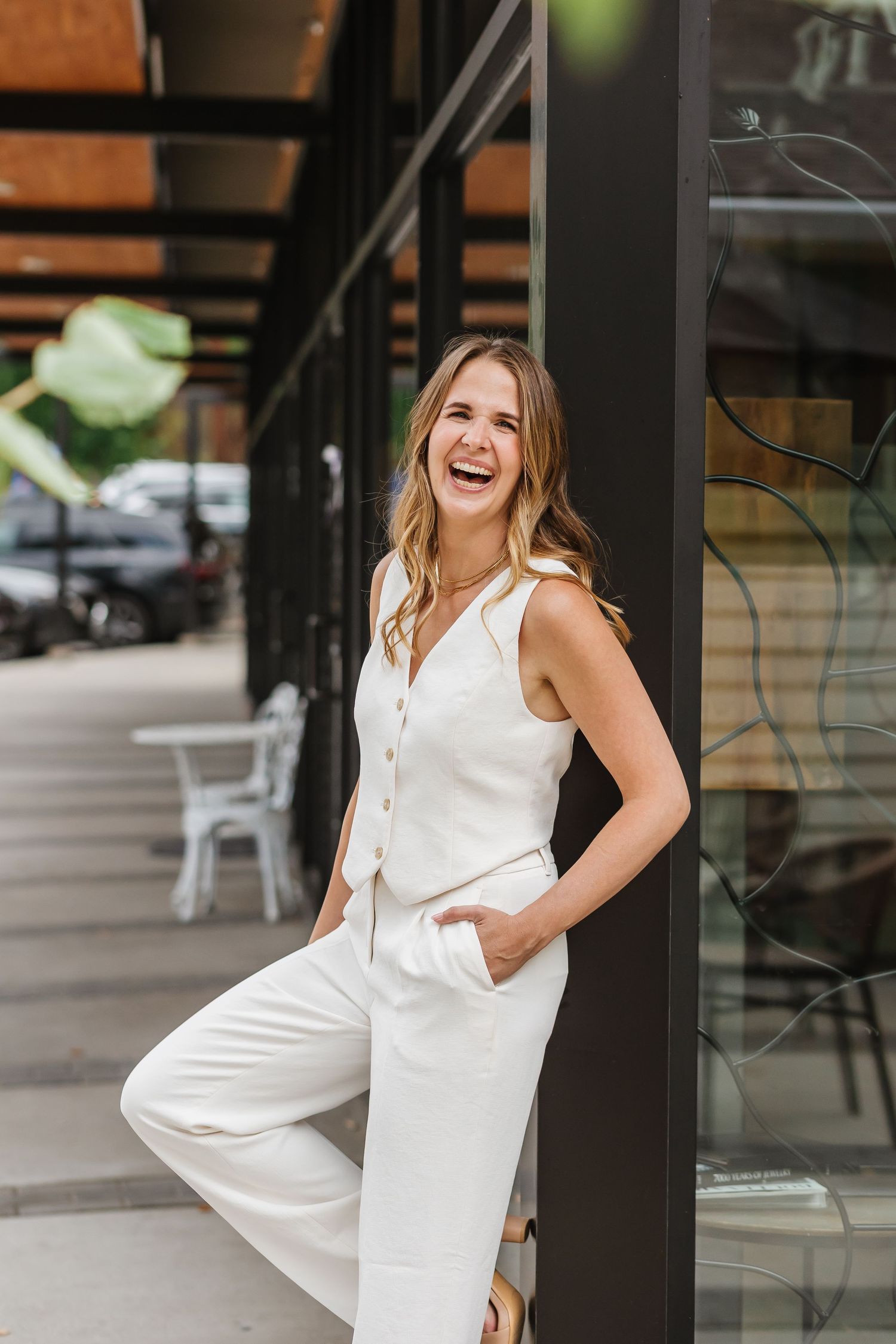 Woman in white sleeveless dress poses playfully against modern architectural backdrop.