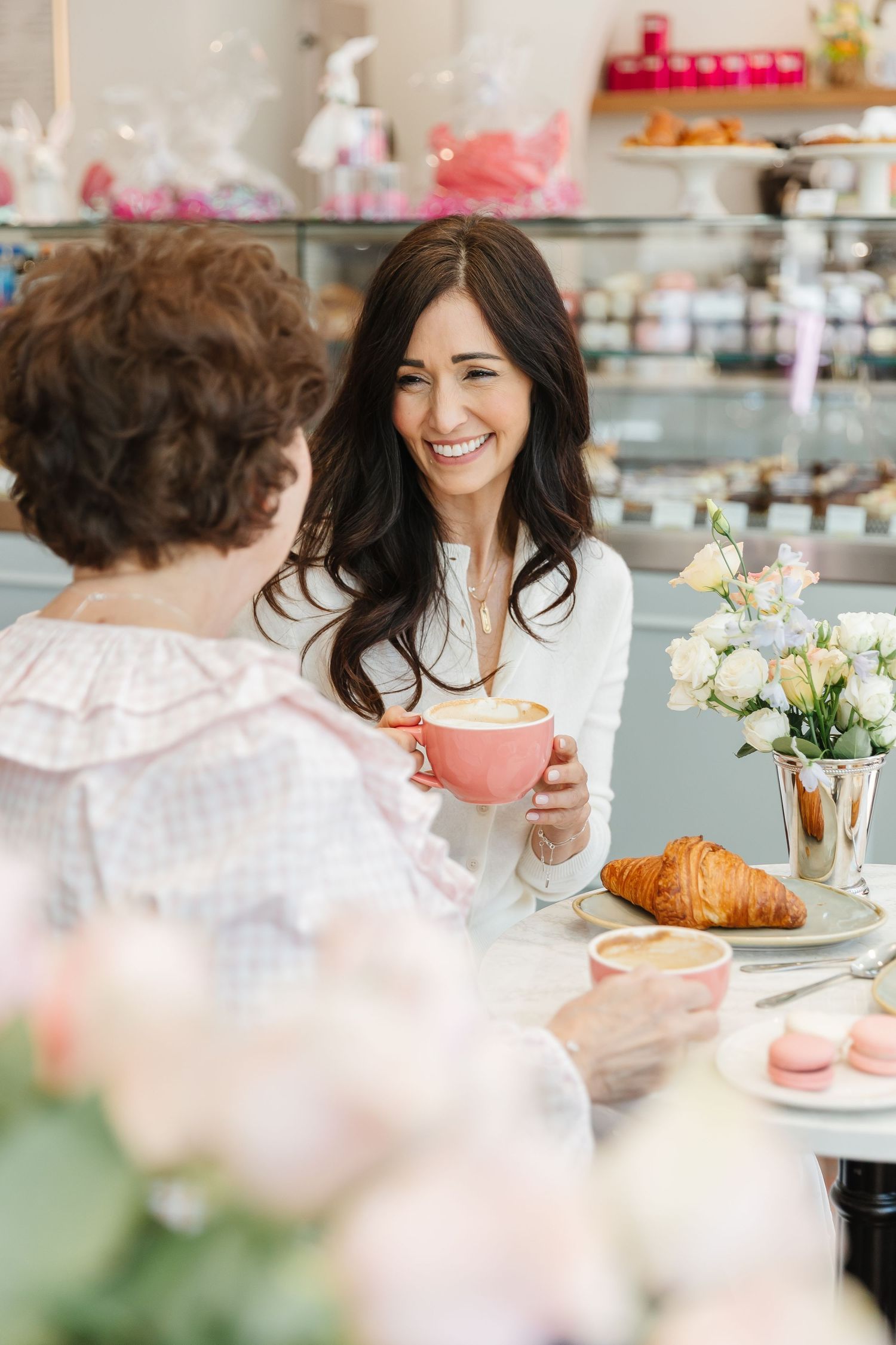Two women laughing and enjoying coffee, croissants, and macarons at a charming pastel-themed French bakery café.