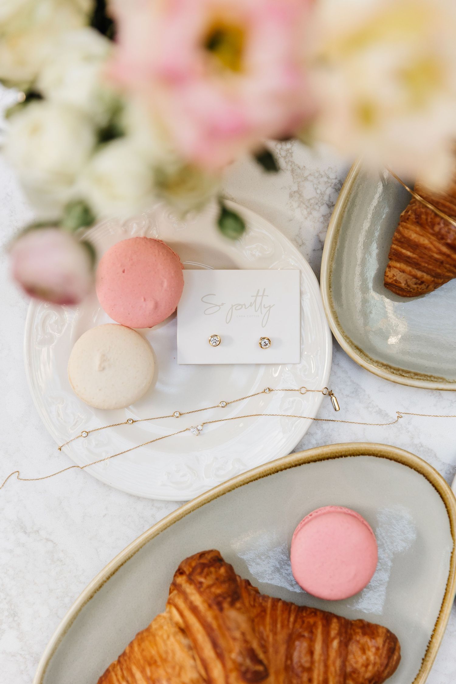 Elegant flat lay of croissants, pastel macarons, gold jewelry, and a sympathy card on a marble table.