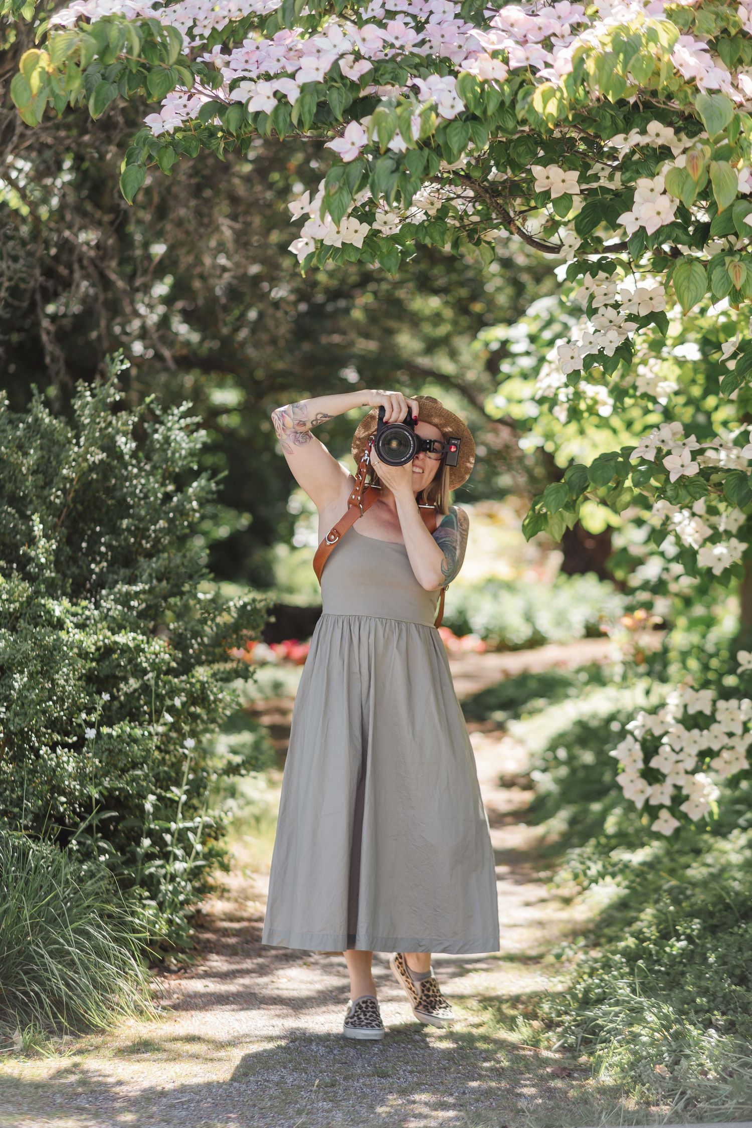 Woman in sage midi dress photographing in a lush garden path beneath flowering dogwood trees.
