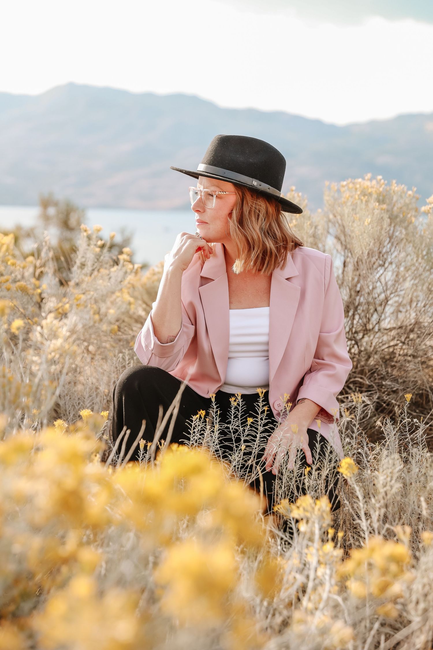 Person in pink blazer and black hat sitting among yellow wildflowers with mountains in background.