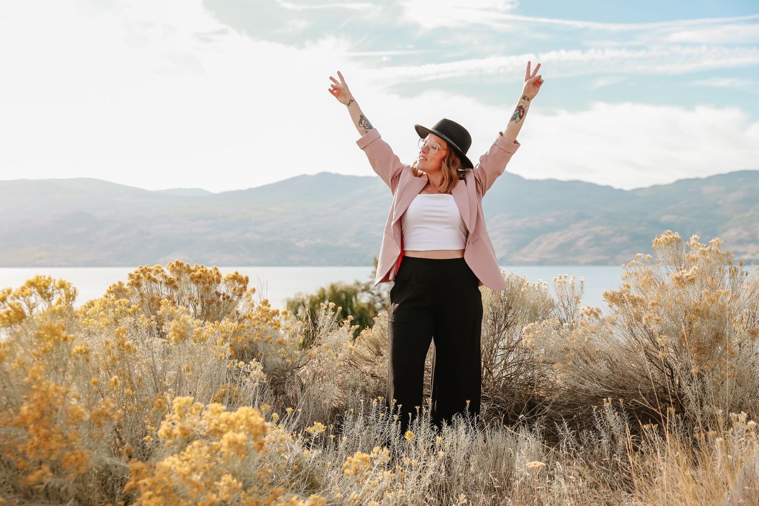 Woman in pink jacket raising arms joyfully in field with mountain landscape.
