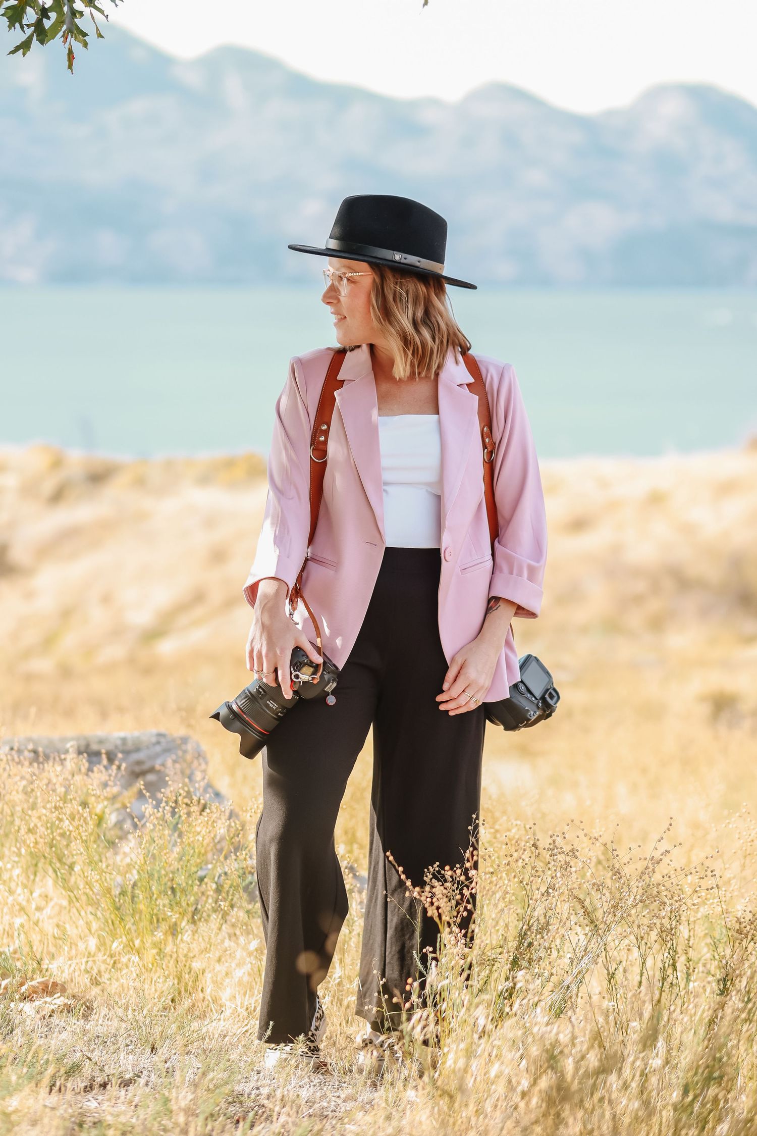 Person wearing pink blazer and black hat walking through tall grass with mountains behind.
