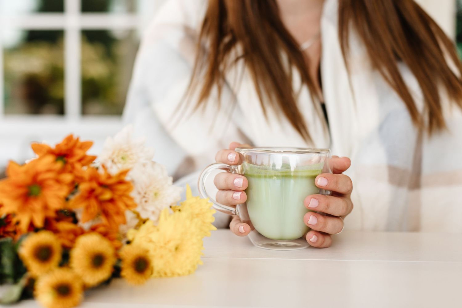 Hands holding beverages with fall decor and sunflowers in a bright setting.