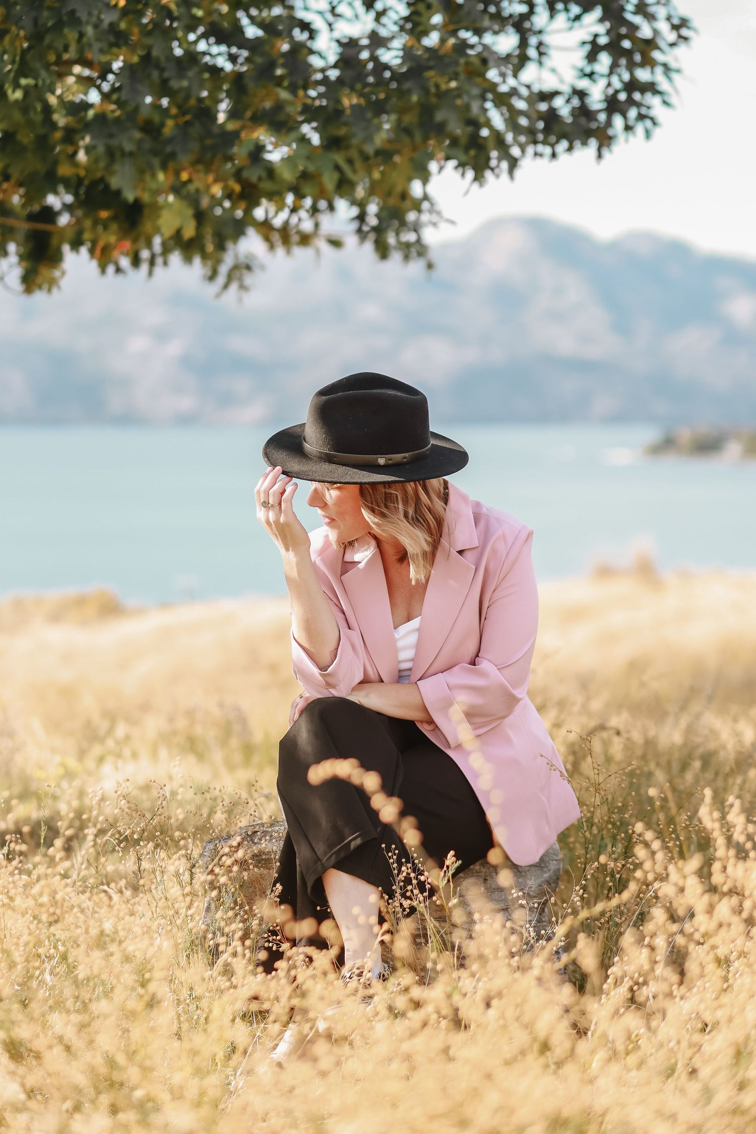 Person in pink blazer and black hat crouching in sunlit meadow with mountains.
