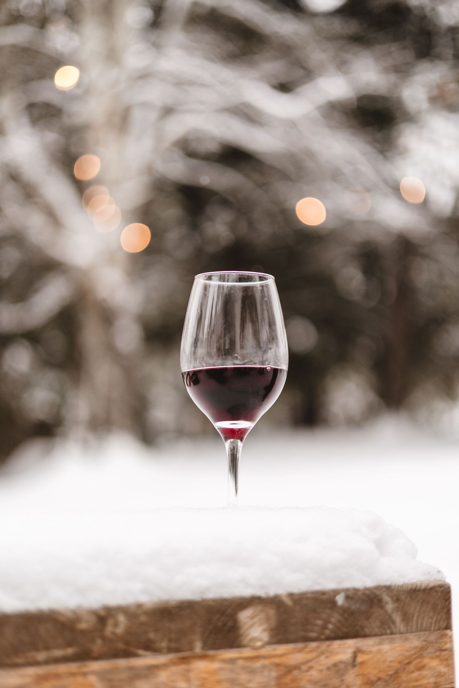Wine glasses with red wine placed on snowy wooden surface with bokeh background.