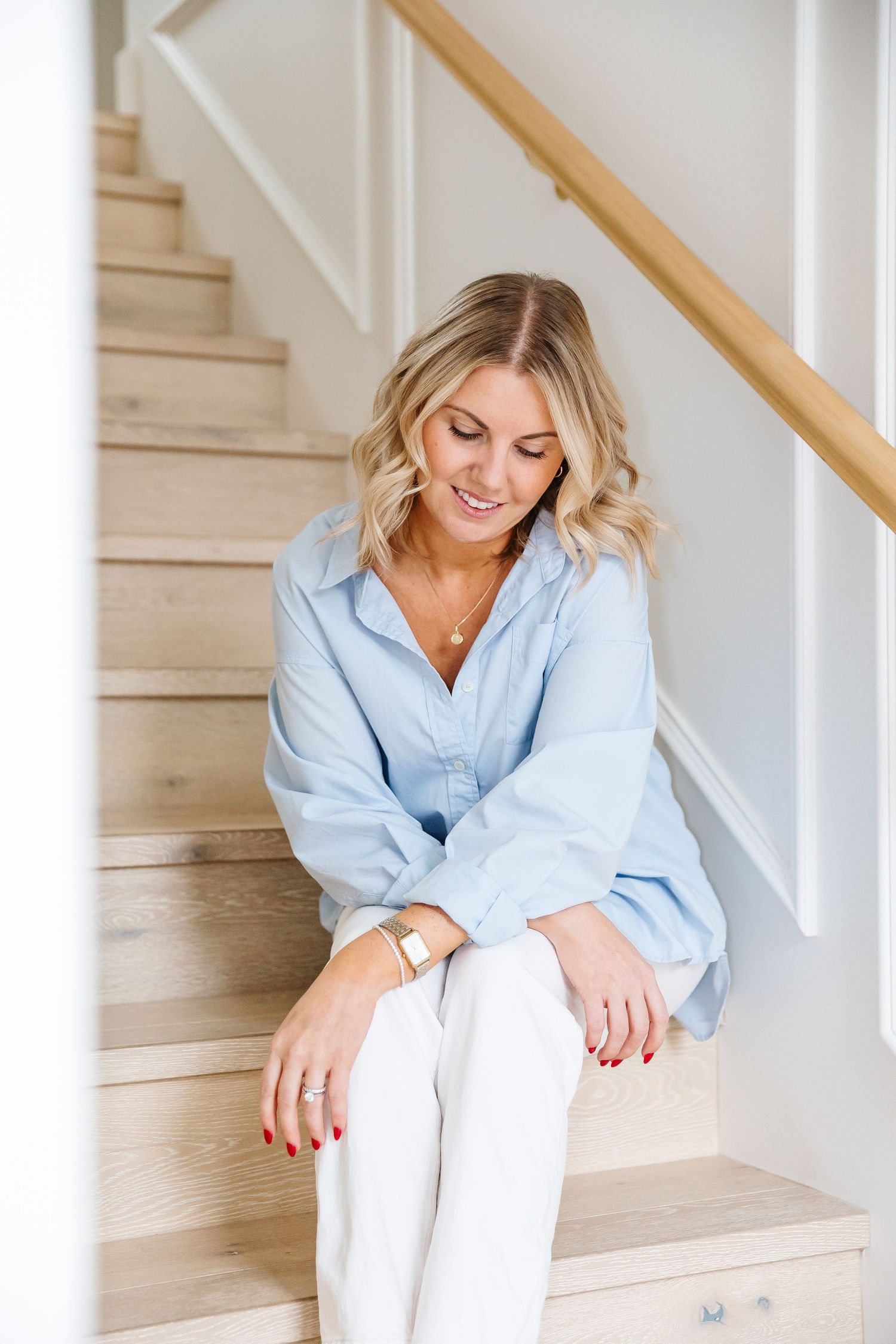 Woman in light blue shirt sitting casually on wooden stairs in bright interior setting.